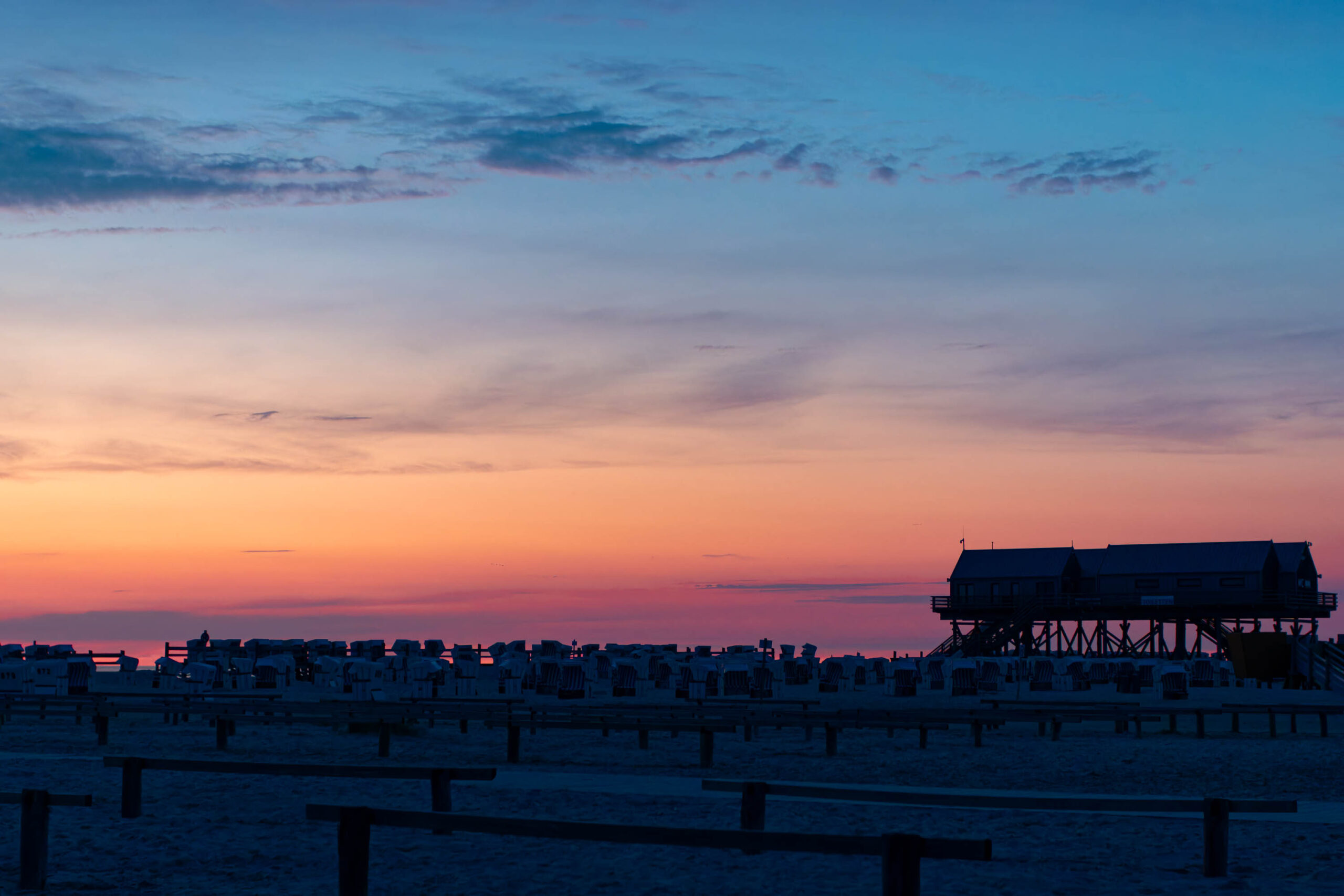 Das Bild zeigt einen Pfahlbau (ein sogenanntes „Strandkorb-Pfahlhaus“) am Ordinger Strand in St. Peter-Ording. Es ist ein typisches Merkmal dieser Küstenregion. Der Himmel ist in einem dramatischen Sonnenuntergang in Rottönen und Violett gefärbt. Die Holzplanken des Strandes sind im Vordergrund deutlich erkennbar. Die Szene vermittelt eine ruhige und friedliche Atmosphäre. Es ist wahrscheinlich ein Sommerabend.