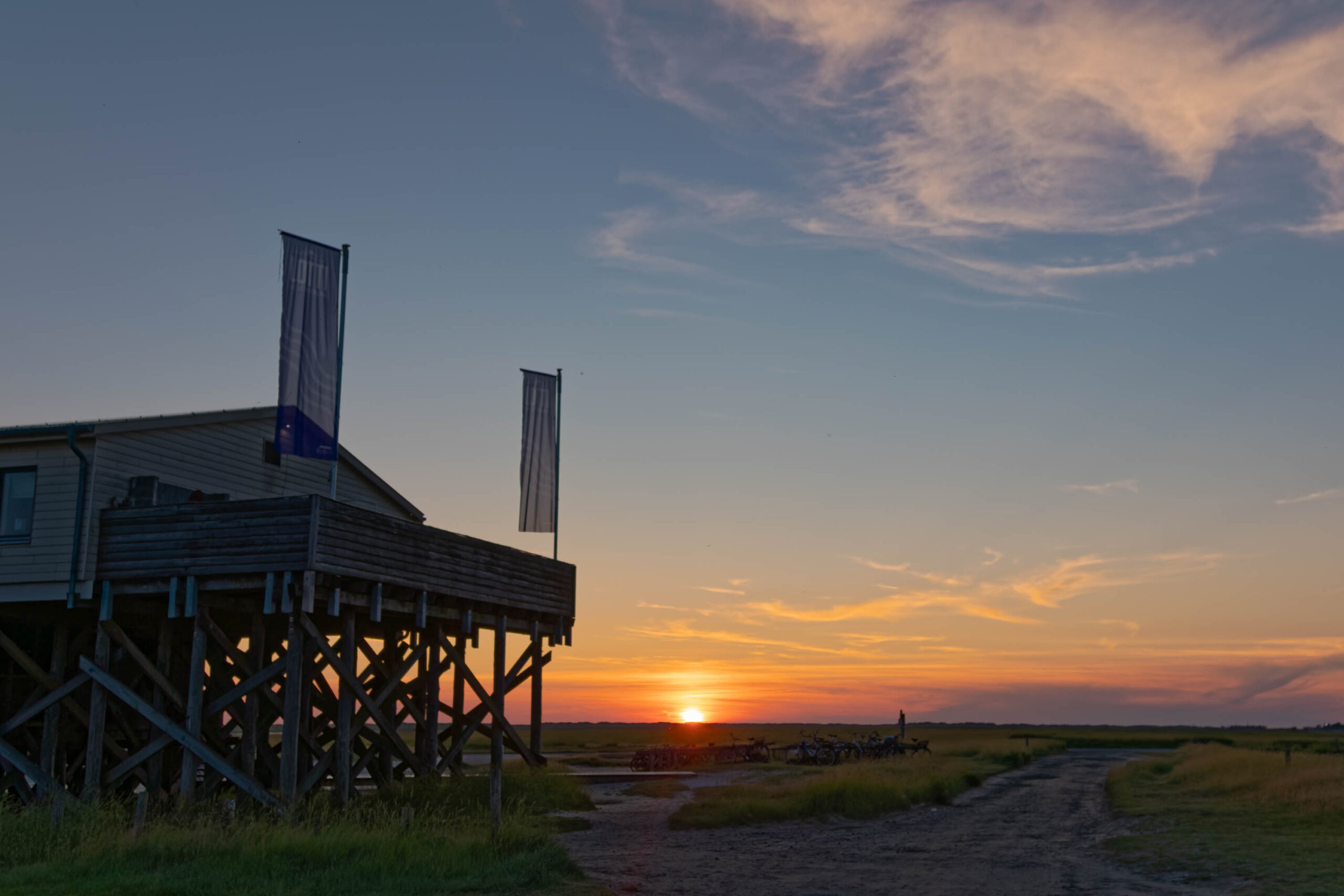 Das Foto zeigt einen Pfahlbau-Restaurantkomplex, der auf Stelzen steht und sich am Strand von St. Peter-Ording befindet. Der Himmel ist in warmen Farben des Sonnenuntergangs gehalten, mit einem orangefarbenen Horizont und einem bläulichen Himmel darüber. Die Sonne geht gerade unter und wirft ein goldenes Licht auf die Szene. Der Strand ist mit Gras bewachsen und ein Feldweg führt in die Ferne. Das Gebäude ist aus Holz und hat mehrere Fenster und eine Terrasse. Zwei Fahnen wehen an den Ecken des Gebäudes. Die Szene vermittelt eine friedliche und entspannte Atmosphäre.