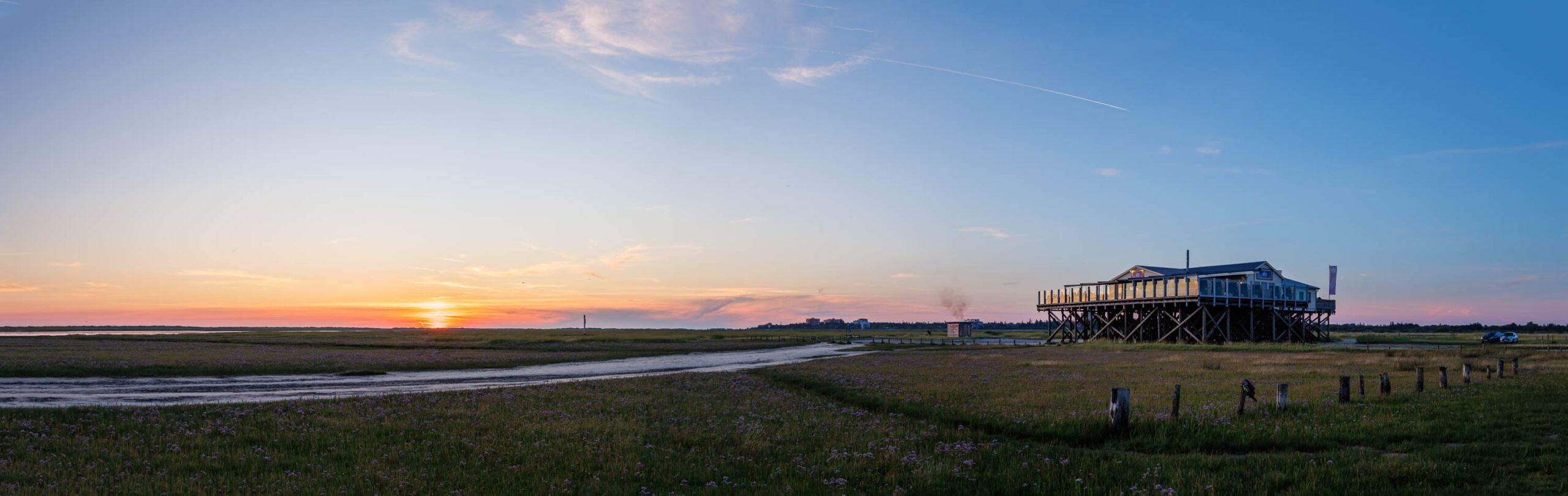 Das Bild zeigt einen Pfahlbau-Restaurantkomplex, bekannt als „Lotti’s“, am Südstrand von St. Peter-Ording, Nordsee. Der Bau steht auf Stelzen inmitten einer grünen Dünenlandschaft. Im Hintergrund ist ein Strandabschnitt mit flachem Wasser und ein weiter Blick über die Nordsee zu erkennen. Der Himmel ist in den Farben eines Sonnenuntergangs gehalten, mit einem sanften Übergang von Blau zu Orange und Rosa. Einige Wolken ziehen über den Himmel. Die Szene vermittelt eine friedliche und entspannte Atmosphäre.