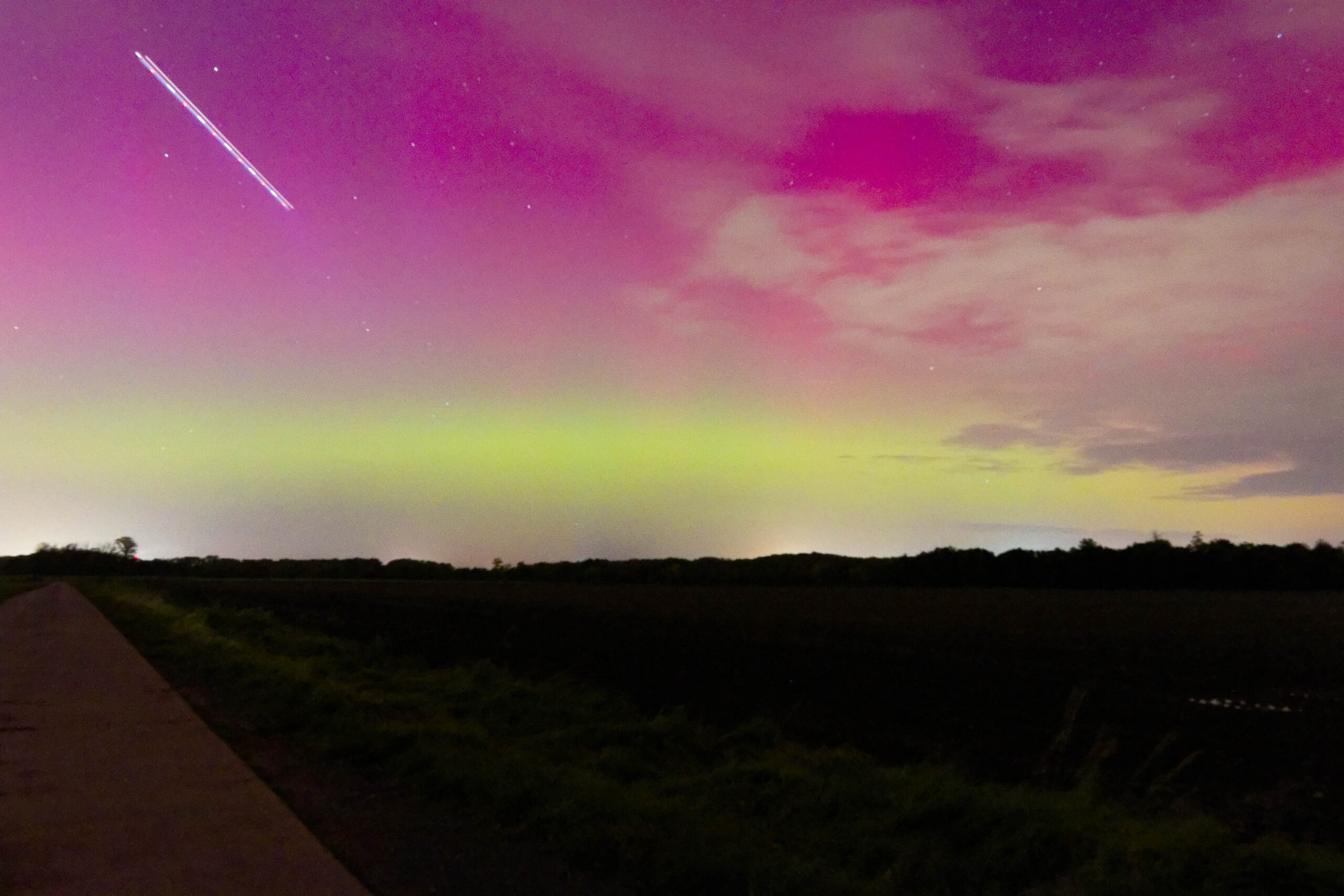 Die Aufnahme zeigt ein beeindruckendes Polarlichter-Phänomen über einer Landschaft in der Region Hannover im Oktober 2024. Der Himmel präsentiert sich in einem faszinierenden Farbspiel aus leuchtendem Rosa und Grün, typisch für Polarlichter. Ein Komet zieht in der oberen Bildhälfte in einer geraden Linie über den Himmel, was dem Bild eine zusätzliche dynamische Komponente verleiht. Im Vordergrund führt ein Feldweg in die Dunkelheit, wobei die Vegetation der Landschaft nur schemenhaft erkennbar ist. Die Sterne am Himmel sind deutlich sichtbar, was auf eine geringe Lichtverschmutzung hindeutet. Die Aufnahme fängt die Magie und die Seltenheit eines solchen Himmelsphänomens ein.
