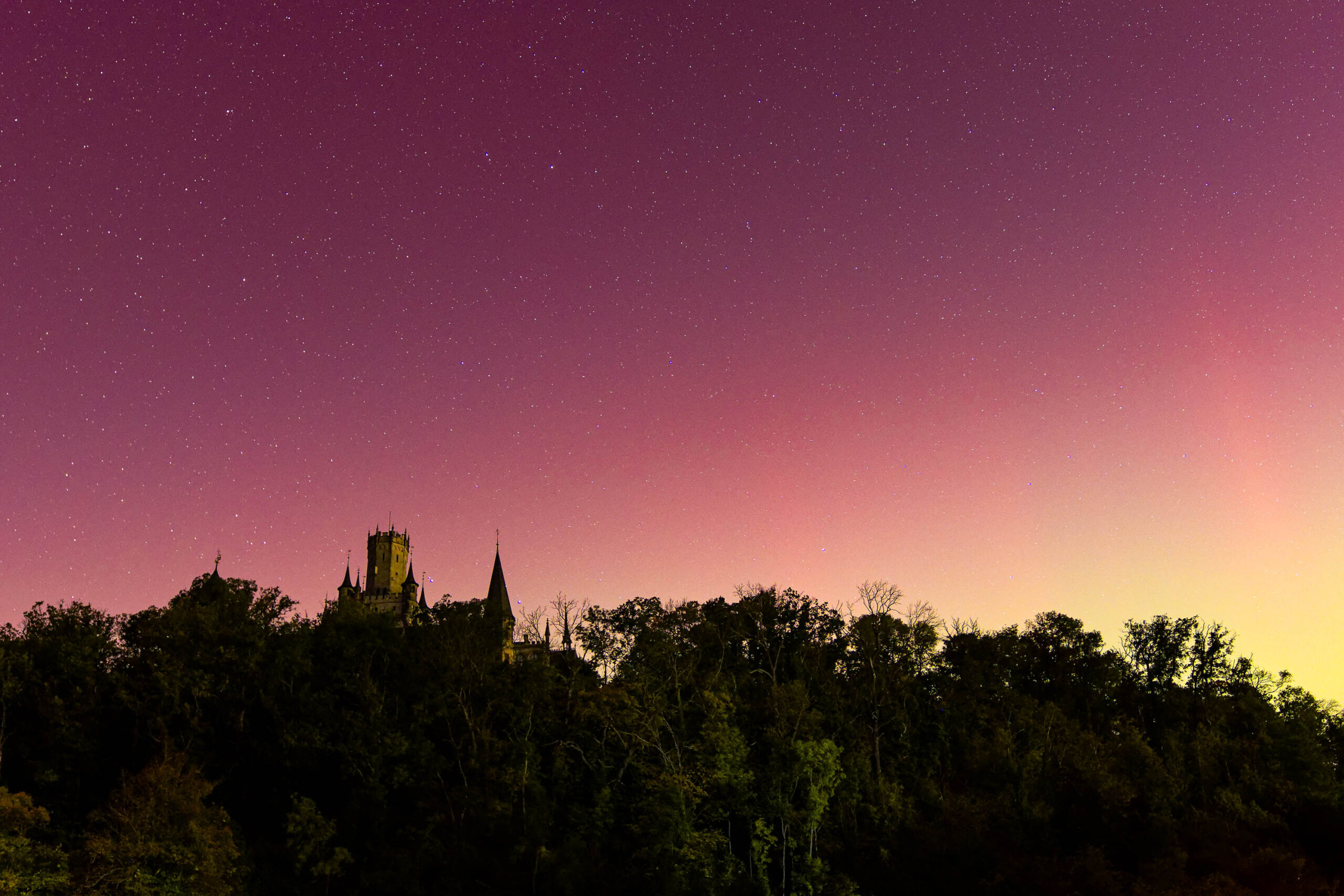 Die Aufnahme zeigt eine nächtliche Szene mit einem markanten Gebäude im Vordergrund, das sich vor einem leuchtenden, violetten Himmel erhebt. Das Gebäude, vermutlich ein historisches Bauwerk, ist silhouettiert und weist mehrere Türme und Zinnen auf. Der Himmel ist von einem intensiven Violettton dominiert, der von einem sanften Orange am Horizont übergeht. Zahlreiche Sterne sind sichtbar, was auf eine klare Nacht und eine geringe Lichtverschmutzung hindeutet. Im Vordergrund befindet sich eine Baumgruppe, die ebenfalls silhouettiert ist und die Szene zusätzlich betont. Die Aufnahme wurde im Oktober 2024 in der Region Hannover aufgenommen und zeigt ein seltenes Phänomen: Polarlichter, die normalerweise in höheren Breitengraden zu sehen sind. Die Aufnahme fängt die Schönheit und das Mysterium dieses seltenen Himmelsphänomens ein.