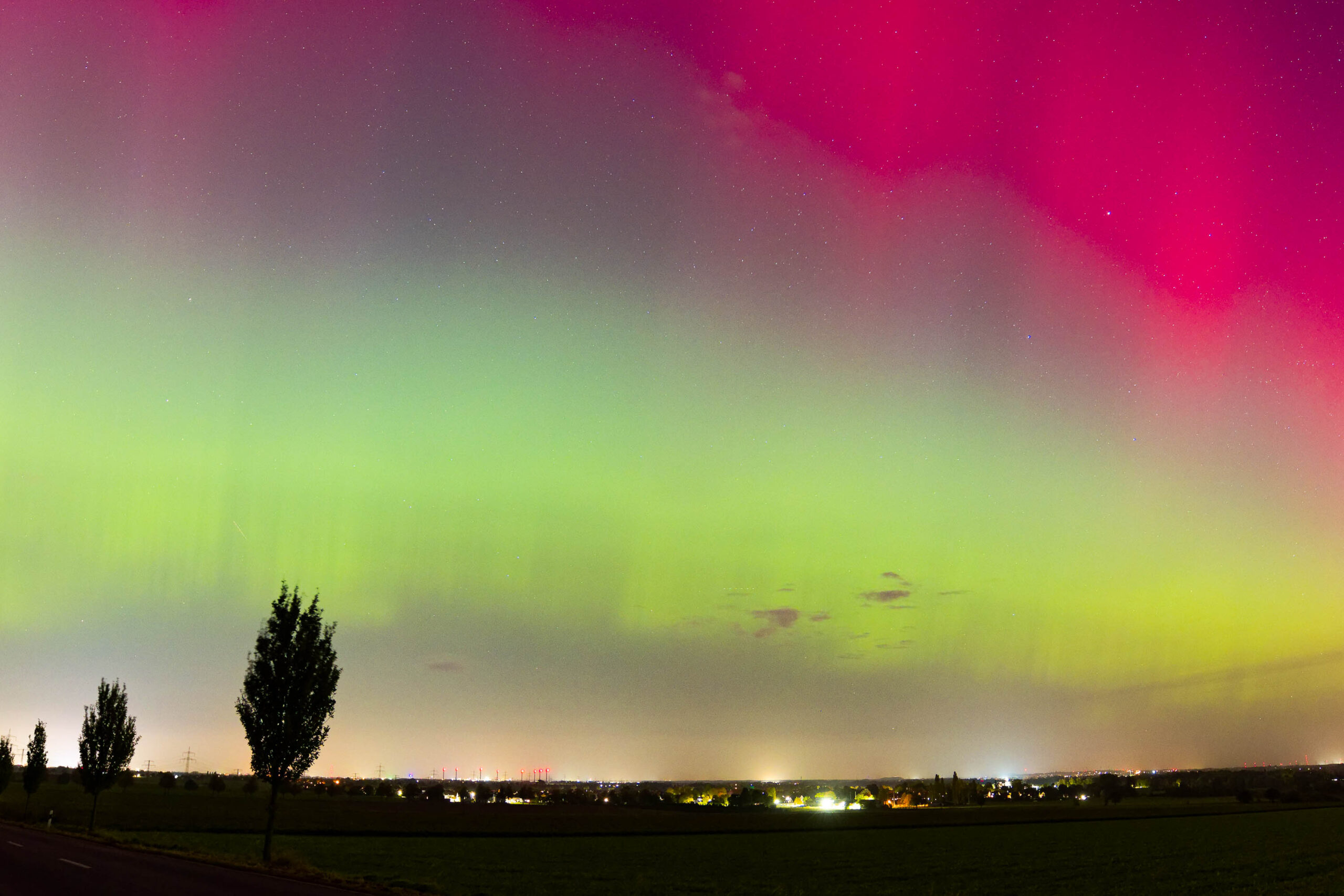 Das Foto zeigt ein beeindruckendes Polarlicht, das den Himmel in leuchtenden Grüntönen und Violett erhellt. Die Szene spielt sich in einer ländlichen Umgebung ab, mit einem grünen Feld im Vordergrund und einem Dorf mit Lichtern am Horizont. Ein einzelner Baum, vermutlich eine Platane, steht im Vordergrund und bildet einen interessanten Kontrapunkt zum leuchtenden Himmel. Die Sterne sind deutlich sichtbar, was auf einen dunklen, klaren Nachthimmel hindeutet. Die Farben des Polarlichts sind intensiv und dynamisch, was die Schönheit und das Phänomen des Polarlichts hervorhebt. Die Aufnahme wurde im Oktober 2024 in der Region Hannover aufgenommen.