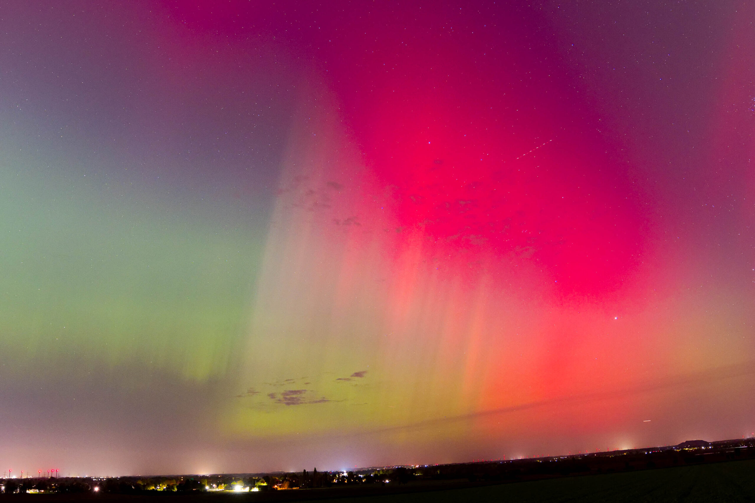 Das Foto zeigt ein beeindruckendes Polarlicht, das sich in den Farben Grün und Pink über einer dunklen Landschaft erstreckt. Der Himmel ist klar und sternenklar, was die Intensität des Polarlichts noch verstärkt. Die Landschaft im Vordergrund ist dunkel und verschwommen, was den Fokus auf das Polarlicht lenkt. Es handelt sich um ein seltenes und faszinierendes Naturschauspiel, das in der Region Hannover im Oktober 2024 beobachtet wurde.