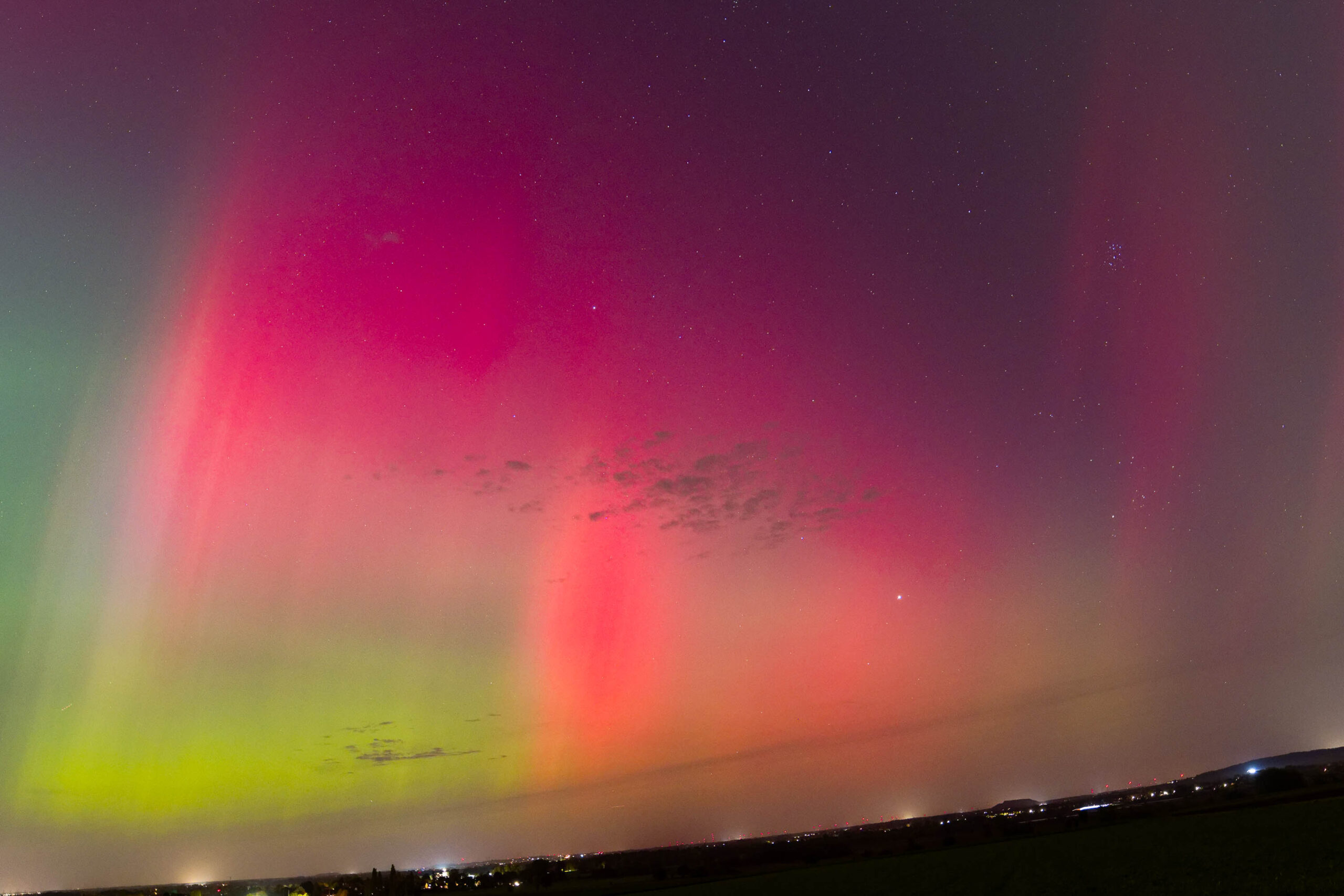 Das Foto zeigt ein beeindruckendes Polarlicht, das den Himmel in leuchtenden Rosa- und Grüntönen erhellt. Die Szene wird durch die sanfte Hügellandschaft im Vordergrund und vereinzelte Lichter am Horizont ergänzt. Der Himmel ist klar und sternenklar, was die Schönheit des Polarlichts noch verstärkt. Die Aufnahme fängt die Magie und das Mysterium des Polarlichts ein und vermittelt ein Gefühl von Ehrfurcht und Staunen.
