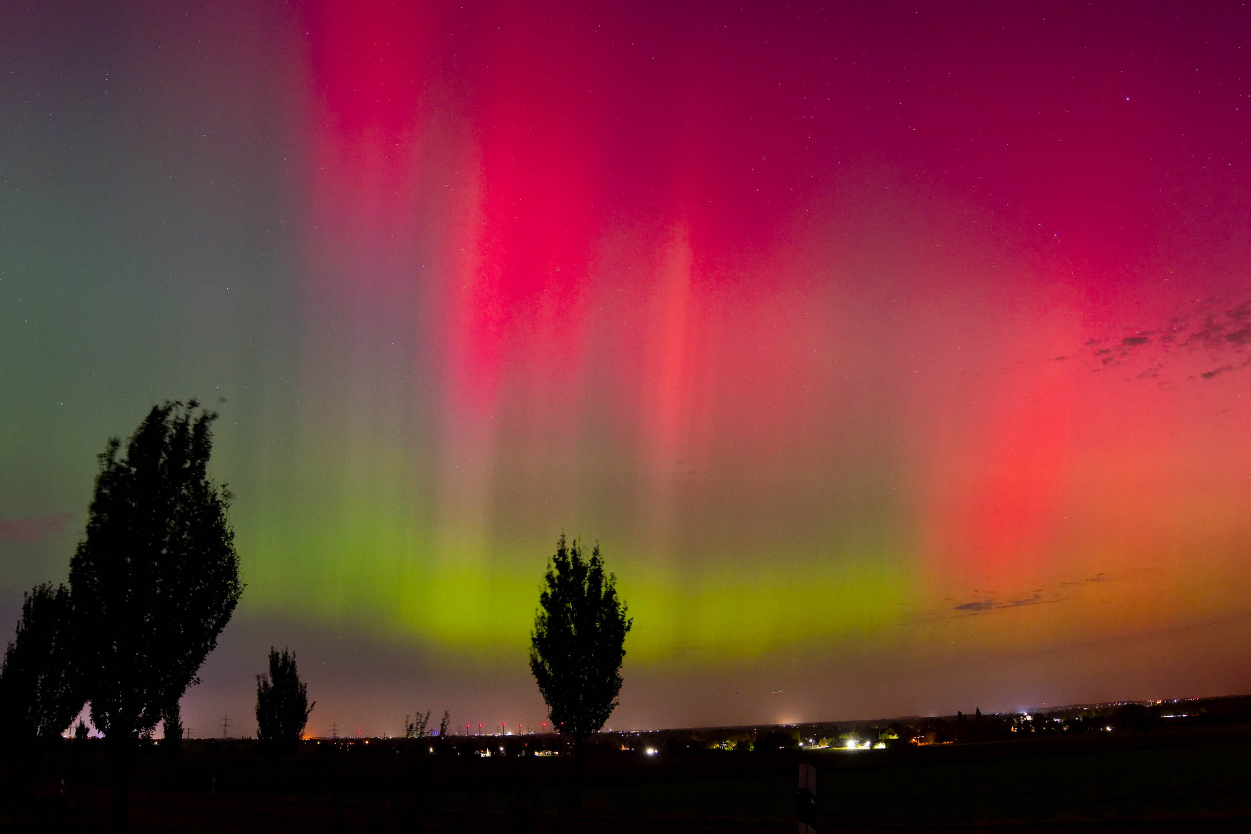 Das Foto zeigt ein beeindruckendes Polarlicht, das sich in leuchtenden Grüntönen und Rottönen über einer Landschaft erstreckt. Der Himmel ist klar und sternenklar, was die Intensität des Polarlichts noch verstärkt. Im Vordergrund stehen einige dunkle Baumgruppen, die die Szene silhouettenartig darstellen. Am Horizont sind vereinzelte Lichter von Siedlungen erkennbar. Die Aufnahme wurde im Oktober 2024 in der Region Hannover aufgenommen.