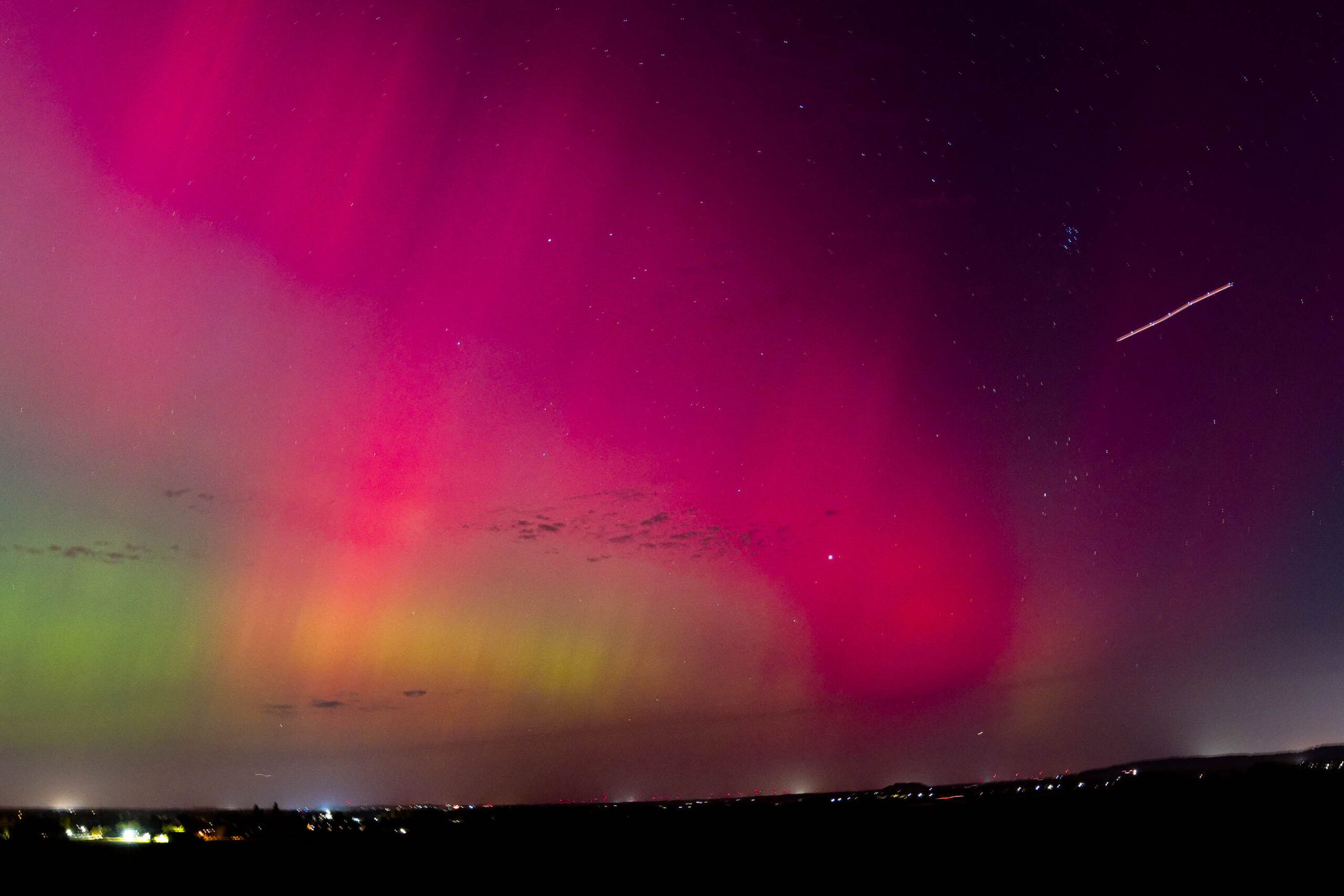 Das Foto fängt die Schönheit und das Mysterium eines Polarlichts ein, das über der Region Hannover im Oktober 2024 erscheint. Die leuchtenden Farben des Polarlichts kontrastieren stark mit der dunklen Landschaft im Vordergrund und erzeugen eine beeindruckende Szene. Ein Komet zieht über den Himmel und fügt dem Bild eine zusätzliche Ebene der Dramatik hinzu.