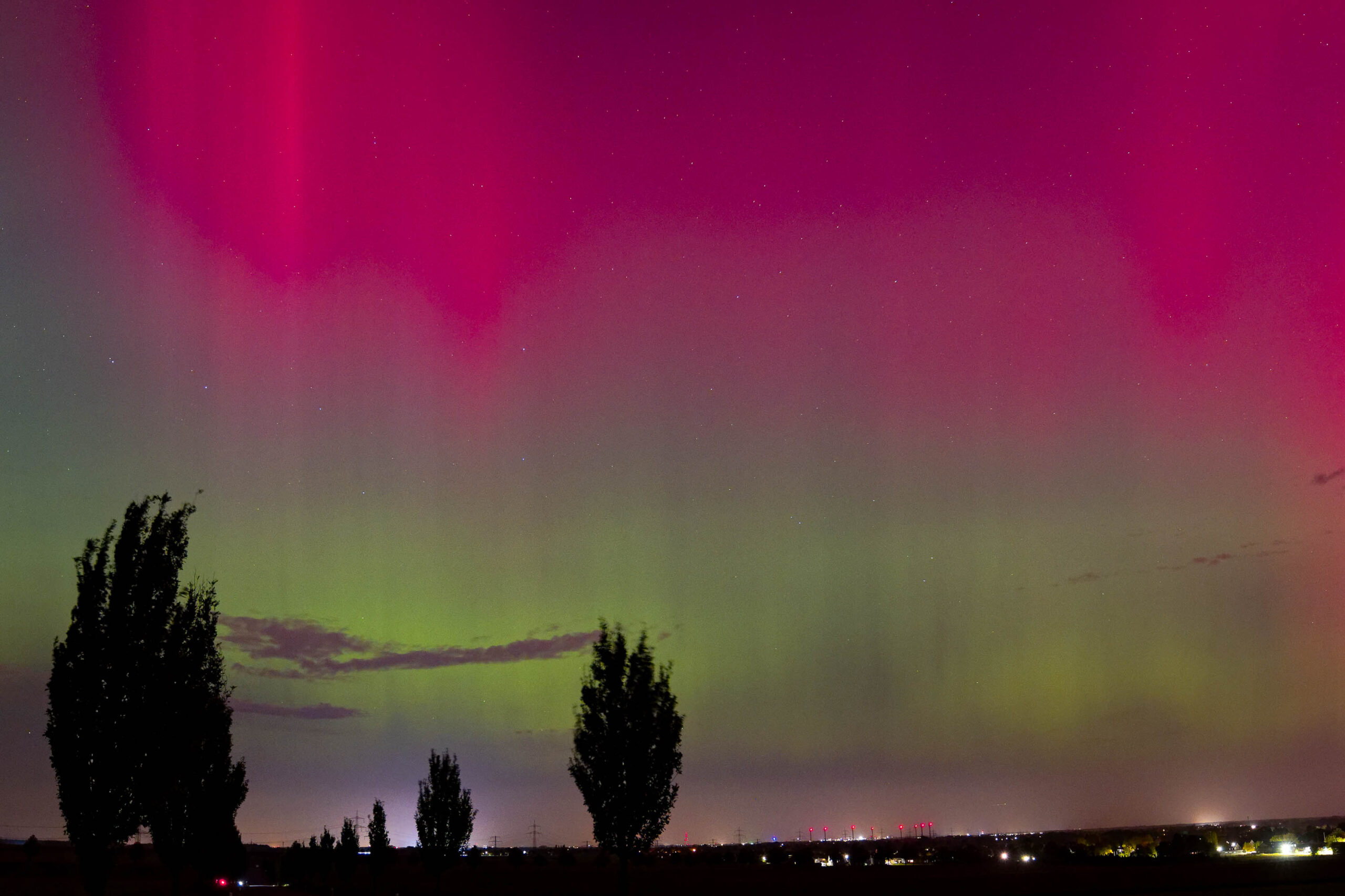 Das Foto fängt ein seltenes und beeindruckendes Polarlichtereignis ein, das über die Landschaft der Region Hannover im Oktober 2024 überzogen ist. Das Polarlicht zeigt sich in leuchtenden Rosa- und Grüntönen, die den Nachthimmel dominieren. Die Silhouette von Bäumen und einer entfernten Stadtlinie bildet einen interessanten Kontrast zu den farbenprächtigen Lichtern. Die Aufnahme vermittelt ein Gefühl von Ehrfurcht und Staunen angesichts der Naturgewalten.