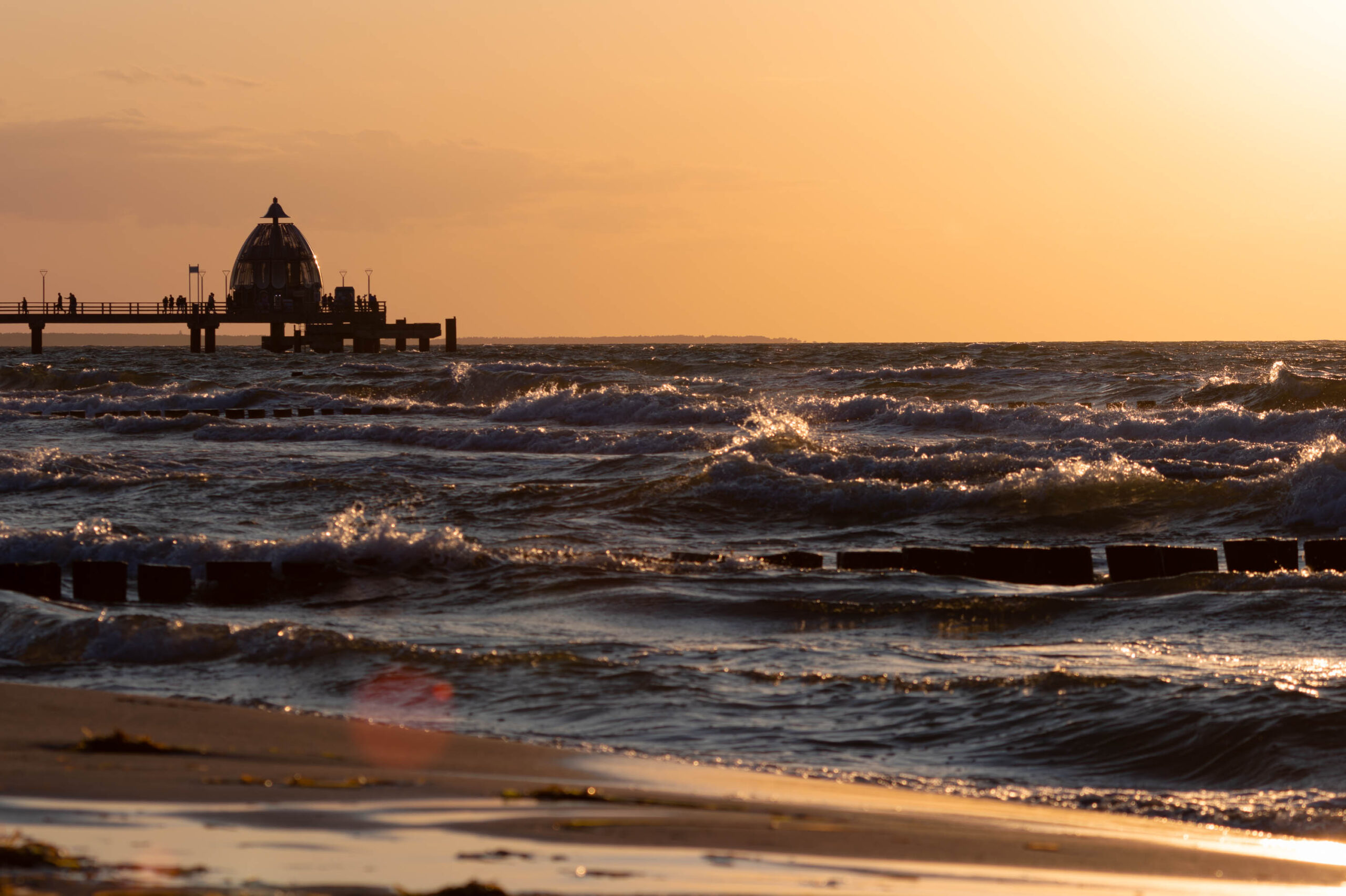 Das Bild zeigt einen Sonnenuntergang an der Küste von Zingst im Sommer 2025. Ein hölzerner Steg erstreckt sich in das Meer hinein. Die Wellen schlagen gegen den Steg und den Strand. Der Himmel ist in warmen Farben, hauptsächlich Orange und Gelb, gefärbt, was eine friedliche und ruhige Atmosphäre schafft. Einige Personen sind auf dem Steg zu sehen, die den Sonnenuntergang genießen. Der Strand ist mit feinem Sand bedeckt, der das Licht reflektiert. Die Szene vermittelt ein Gefühl von Ruhe und Schönheit der Natur.