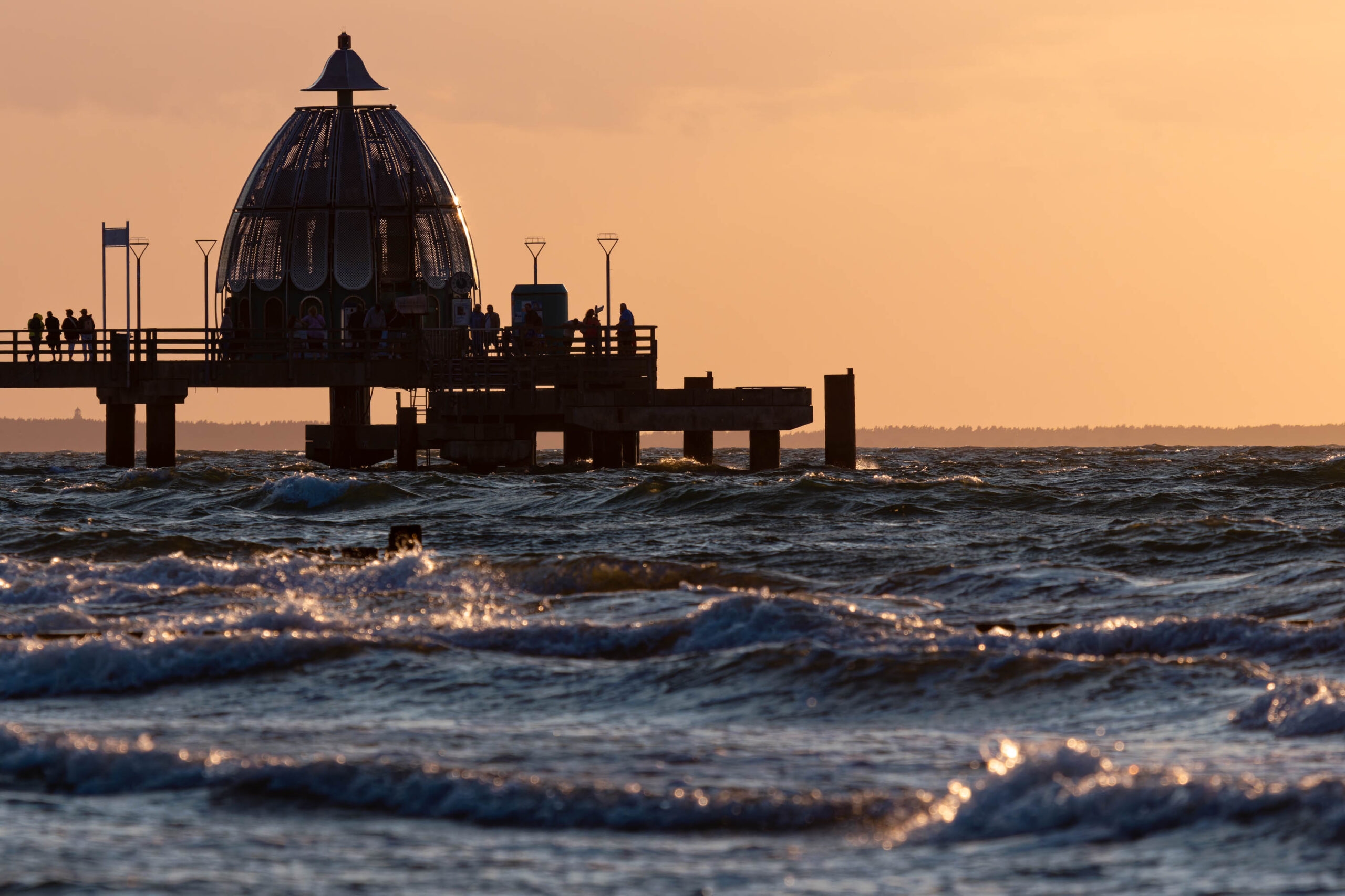 Das Bild zeigt einen Steg, der sich in das Meer erstreckt, wobei der Himmel in warmen Orange- und Gelbtönen leuchtet. Das Wasser ist leicht aufgewühlt und reflektiert das Licht. Im Hintergrund sind einige Gebäude und Bäume zu sehen. Die Szene vermittelt eine friedliche und entspannte Atmosphäre.