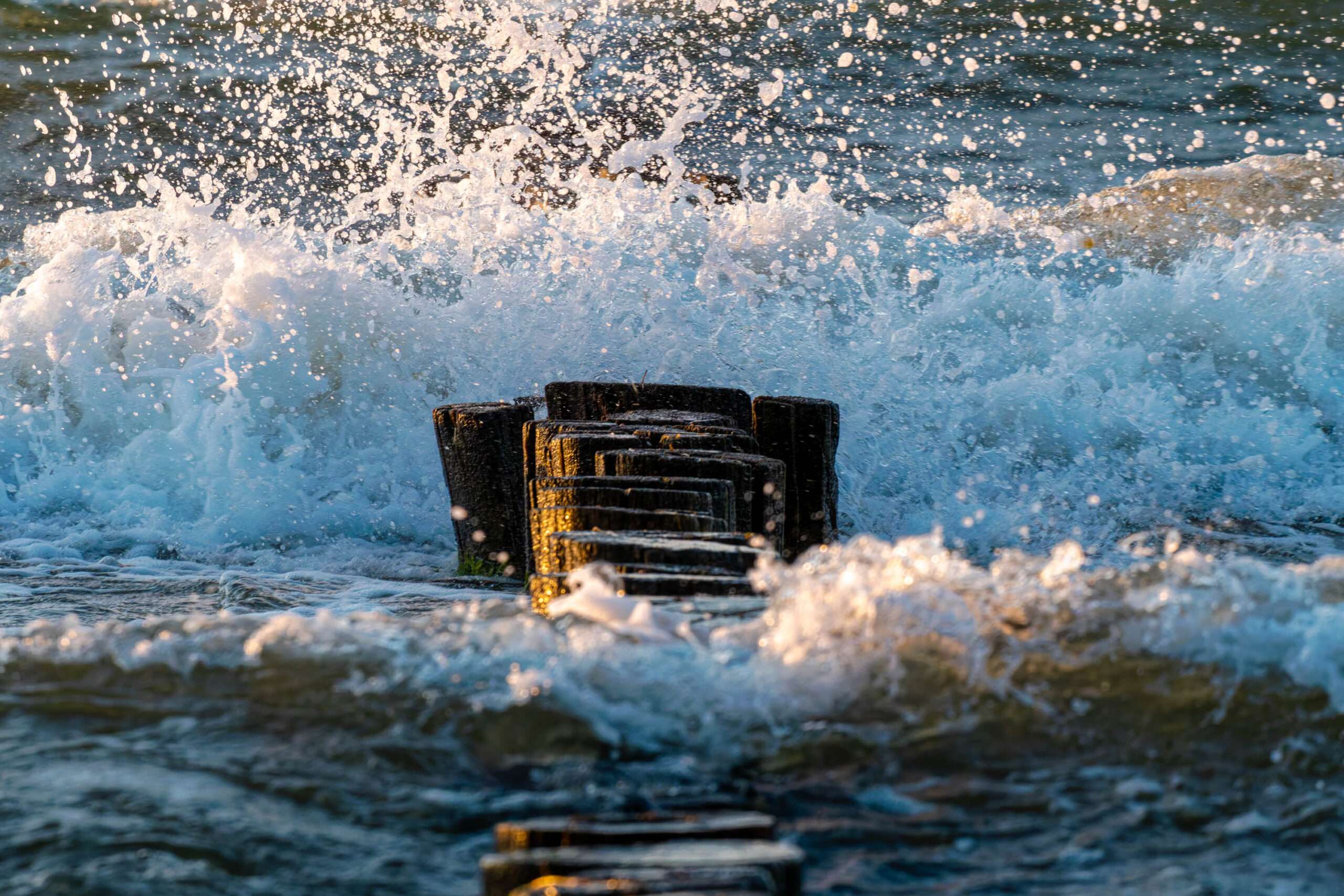 Das Bild zeigt einen verwitterten Holzpfahl, der aus dem Wasser ragt und von starken Wellen umspült wird. Das Wasser ist in Bewegung und erzeugt eine dynamische Szene. Die Sonne scheint durch die Wolken und beleuchtet das Wasser und den Pfahl. Das Bild vermittelt ein Gefühl von Kraft und Bewegung.