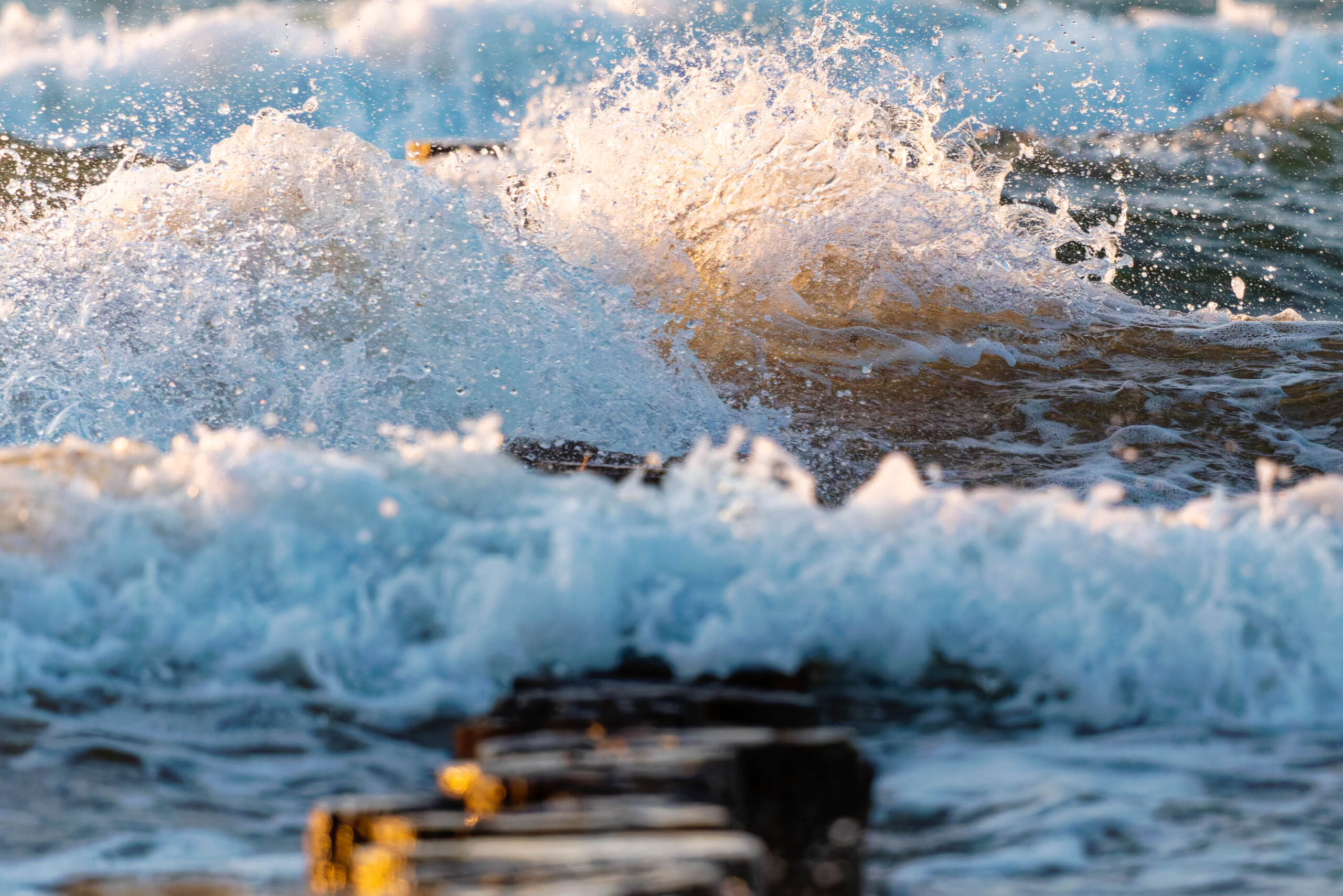 Das Bild zeigt eine dynamische Nahaufnahme von Wellen, die mit Kraft gegen eine Holzkonstruktion, vermutlich einen Steg oder eine Mole, schlagen. Das Wasser ist in verschiedenen Blautönen dargestellt, wobei die Gischt und die aufspritzenden Wassertropfen in einem goldenen Licht schimmern. Die Textur des Holzes ist deutlich erkennbar und kontrastiert mit der fließenden Bewegung des Wassers. Die Szene vermittelt ein Gefühl von Kraft und Bewegung, während die goldenen Reflexionen eine warme Atmosphäre schaffen. Der Fokus liegt auf dem Moment des Aufpralls, der die Energie der See verdeutlicht. Der Kontext 'Zingst, Sommer 2025' deutet auf eine Urlaubslandschaft hin, in der die Naturgewalt der See im Vordergrund steht.