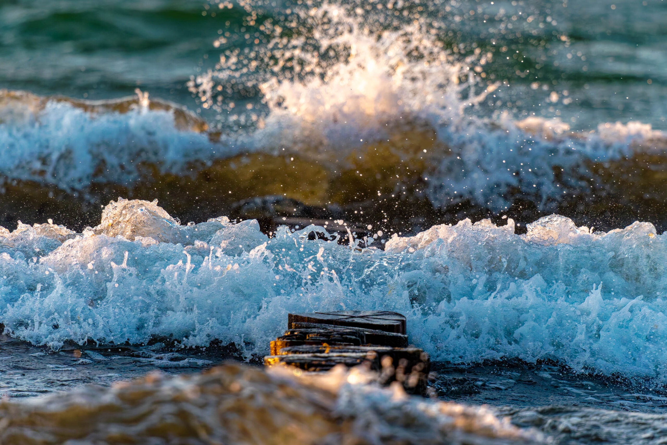Das Foto zeigt einen Holzsteg, der von Wellen überflutet wird. Das Wasser ist blau und türkis und die Wellen sind weiß und schaumig. Der Himmel ist bewölkt. Das Foto wurde wahrscheinlich an einem stürmischen Tag aufgenommen. Die Szene vermittelt ein Gefühl von Bewegung und Kraft.