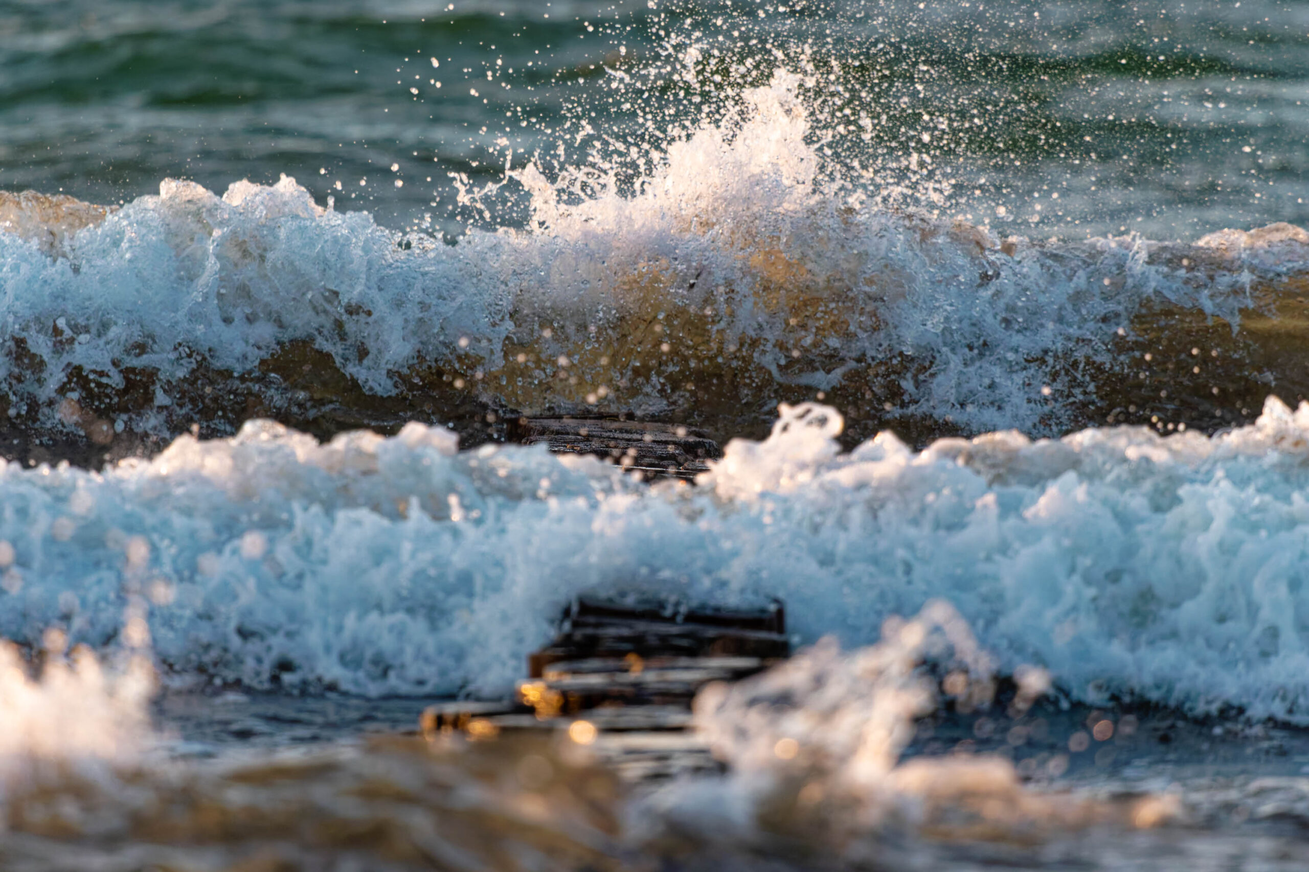 Eine dynamische Nahaufnahme zeigt Wellen, die mit Kraft gegen einen hölzernen Steg schlagen. Das Wasser ist in verschiedenen Blautönen dargestellt, wobei die Wellen weiß schäumen und Gischt in die Luft schleudern. Der Steg selbst ist dunkel und strukturiert, was einen Kontrast zum schäumenden Wasser bildet. Die Szene vermittelt ein Gefühl von Bewegung und Energie.