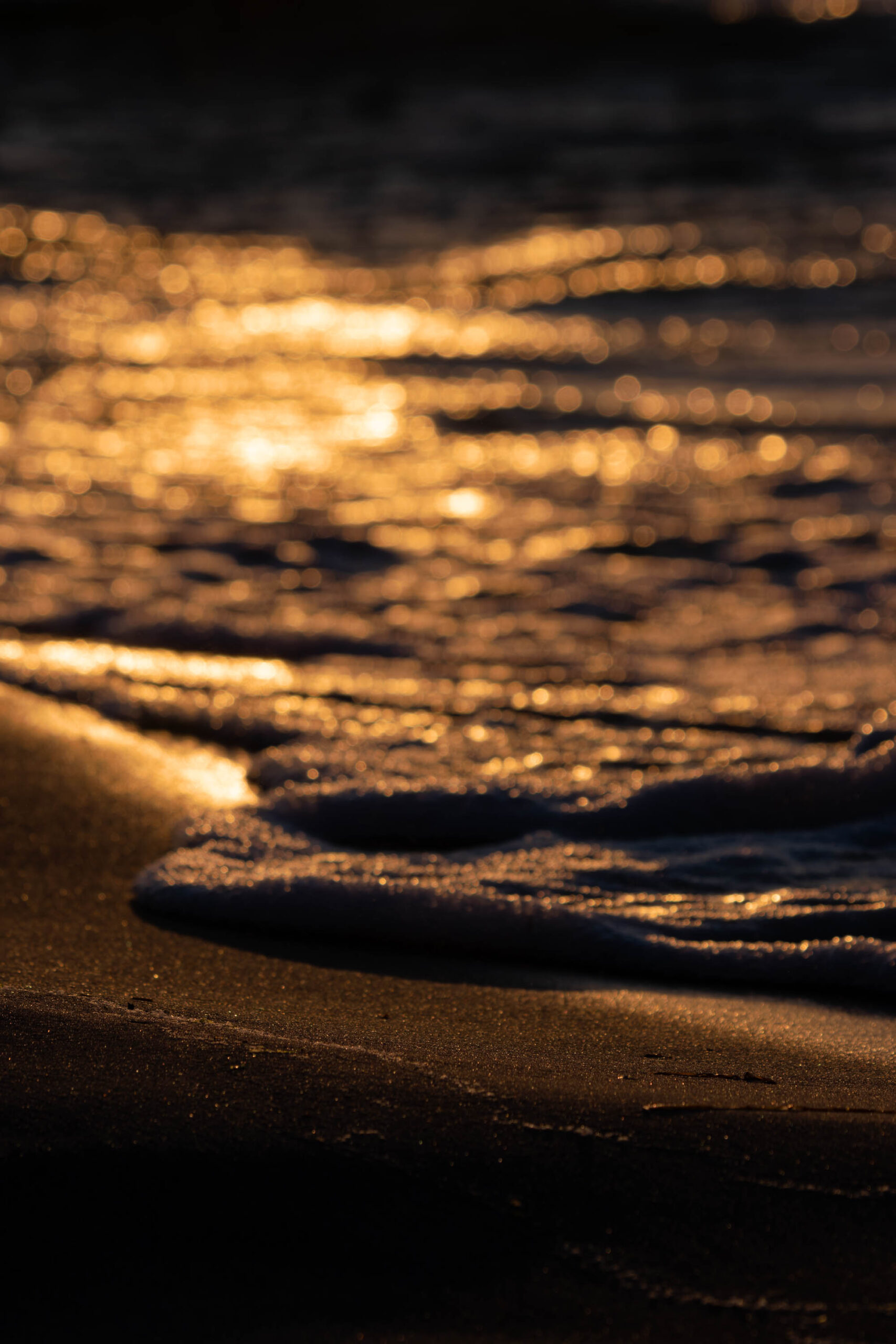 Das Bild zeigt einen Strand bei Sonnenuntergang oder Sonnenaufgang. Der Sand im Vordergrund ist golden und reflektiert das Licht. Wellen brechen sanft am Ufer. Der Himmel ist von Wolken bedeckt, die ebenfalls das Licht reflektieren und einen goldenen Schein erzeugen. Die Szene vermittelt eine friedliche und ruhige Atmosphäre.