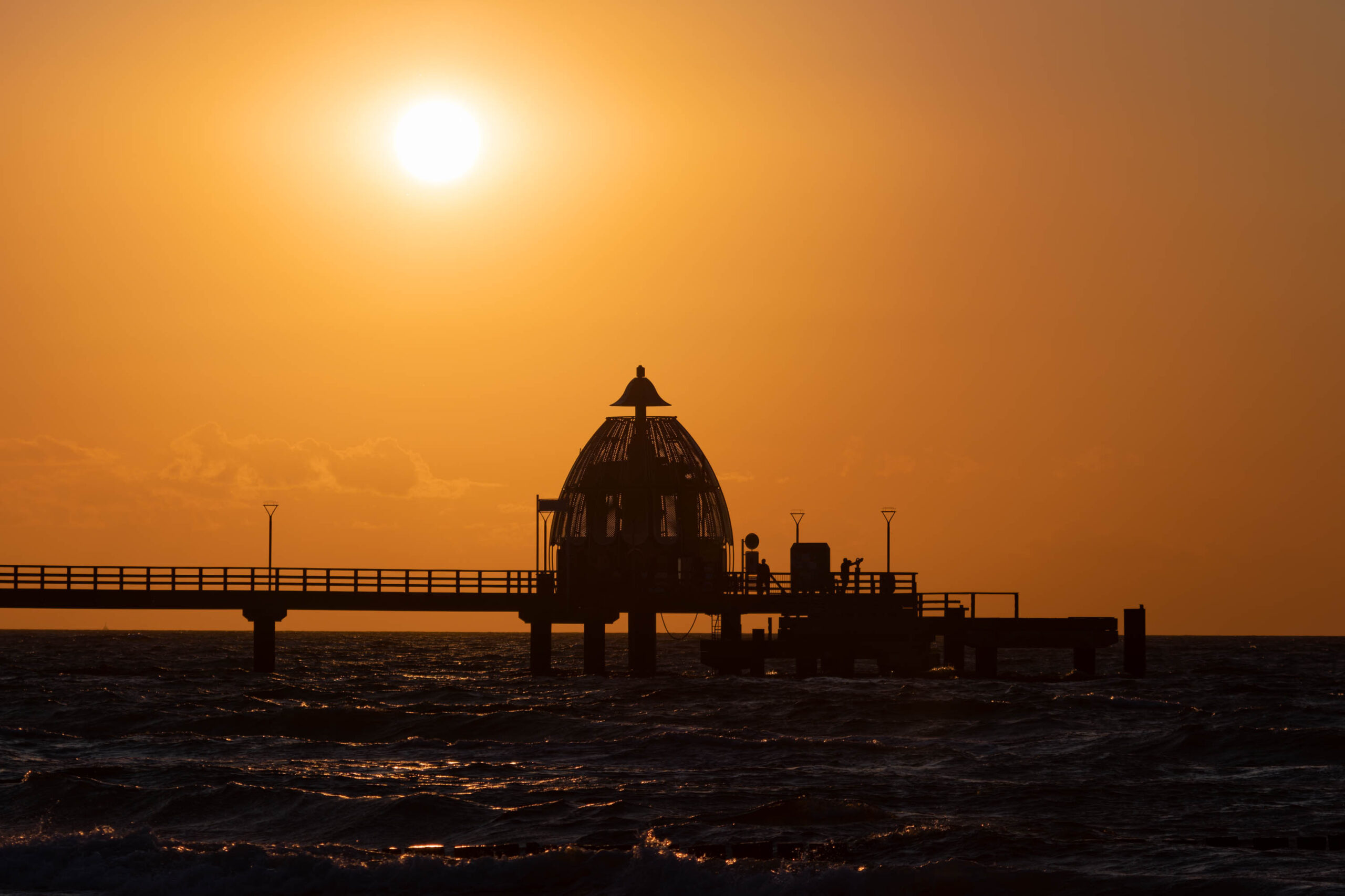 Das Bild zeigt eine Silhouette einer hölzernen Seebrücke vor einem dramatischen, orangefarbenen Sonnenuntergang. Die Seebrücke erstreckt sich horizontal über das Bild, wobei die Struktur durch ihre dunkle Kontur deutlich hervortritt. Die Brücke scheint auf Pfählen zu stehen, die ins Wasser führen. Ein kleines, kuppelartiges Gebäude befindet sich mittig auf der Brücke. Einige Personen sind auf der Brücke zu erkennen, die sich in Silhouette abzeichnen. Der Himmel ist in einem intensiven Orange-Ton gehalten, was eine warme und stimmungsvolle Atmosphäre erzeugt. Das Wasser ist leicht wellig und spiegelt das Licht des Sonnenuntergangs wider. Die Szene vermittelt ein Gefühl von Ruhe und Frieden.