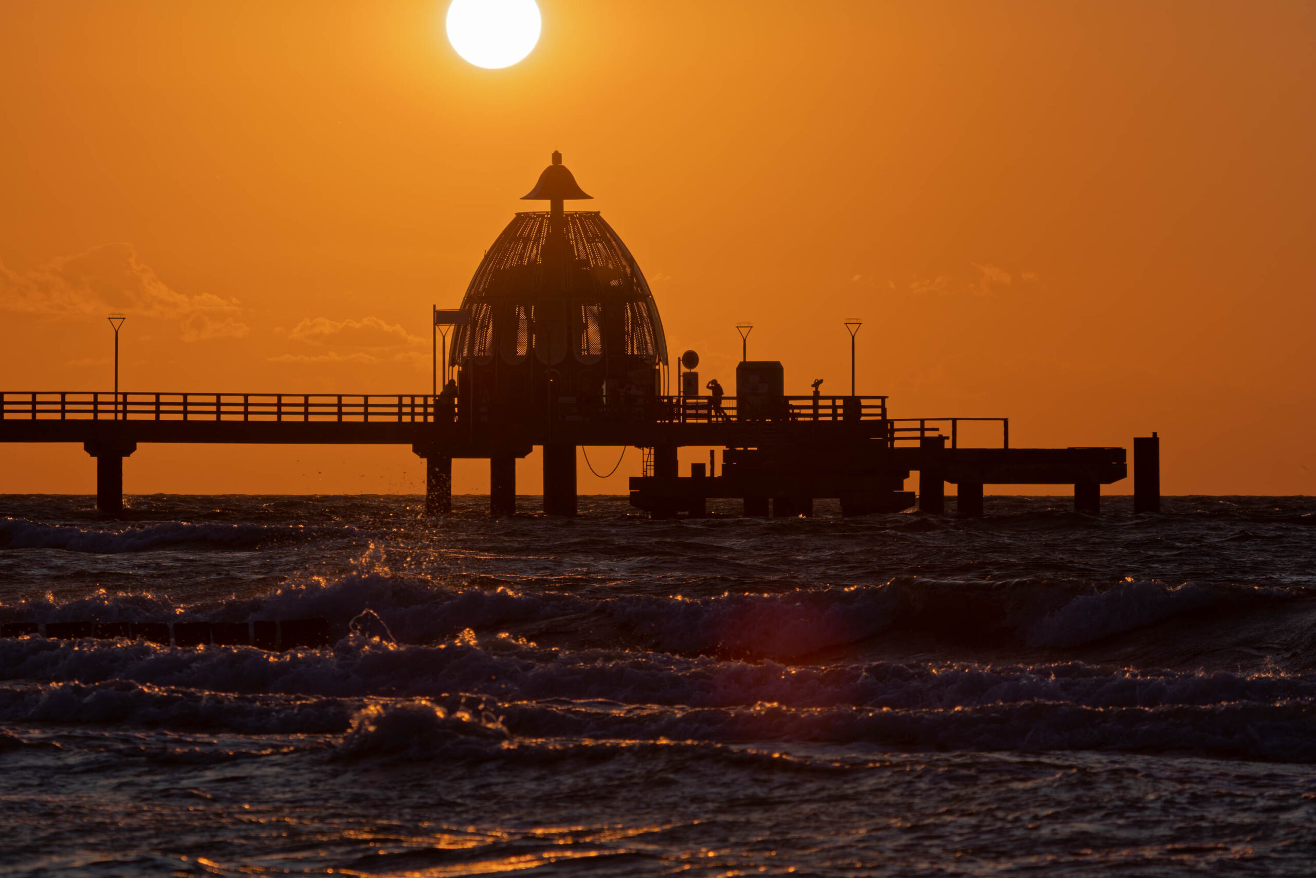 Die Aufnahme zeigt einen Pier, der sich in den Sonnenuntergang erstreckt. Der Pier ist von Menschen belebt, die sich auf der Promenade aufhalten und die Aussicht genießen. Der Himmel ist in warmen Orange- und Rottönen gehalten, die sich im Wasser spiegeln. Die Silhouette des Piers und der Menschen wird gegen das helle Licht des Sonnenuntergangs hervorgehoben. Die Aufnahme vermittelt eine friedliche und entspannte Atmosphäre.
