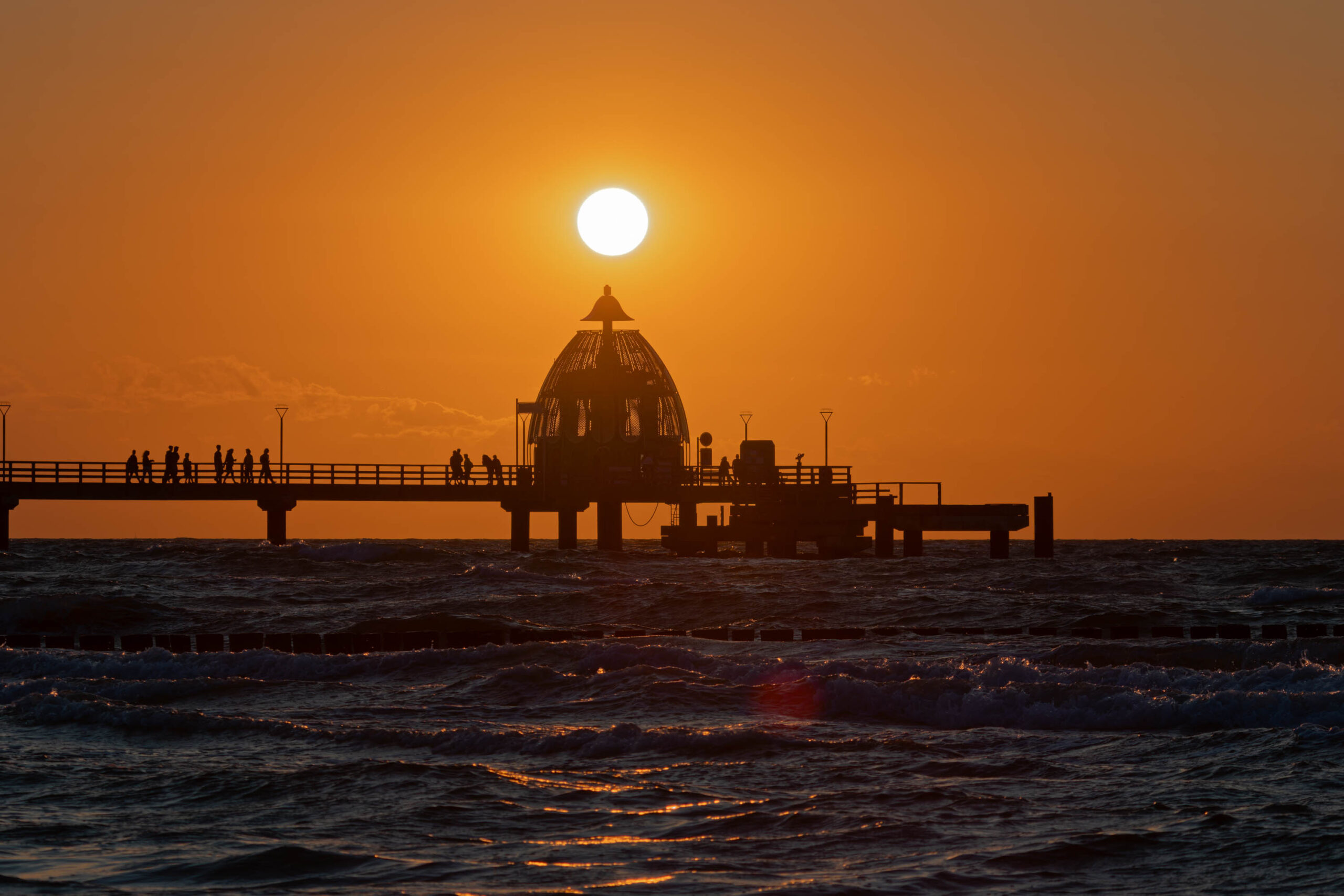 Das Bild zeigt einen Steg, der sich in den Sonnenuntergang erstreckt. Die Silhouette von Menschen ist auf dem Steg erkennbar. Der Himmel ist in warmen Orange- und Rottönen gehalten, was eine stimmungsvolle Atmosphäre schafft. Das Wasser ist leicht wellig und reflektiert das Licht des Sonnenuntergangs. Die Szene vermittelt ein Gefühl von Ruhe und Frieden.