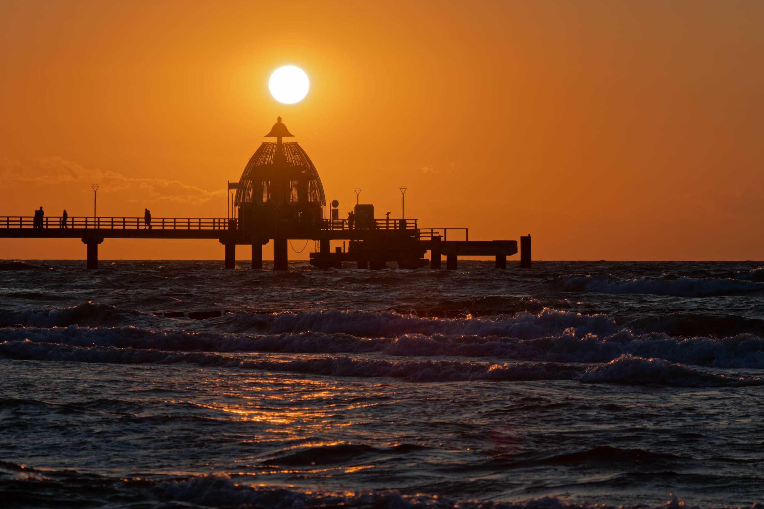 Das Bild zeigt einen Sonnenuntergang über dem Meer, wobei ein Steg im Vordergrund steht. Der Himmel ist in leuchtenden Orange- und Rottönen gehalten, die sich im glitzernden Wasser spiegeln. Mehrere Personen befinden sich auf dem Steg, die silhouettiert vor dem Sonnenuntergang stehen. Die Szene vermittelt eine friedliche und ruhige Atmosphäre. Der Steg scheint ein markantes Merkmal der Küstenlinie zu sein, möglicherweise ein Wahrzeichen der Region.