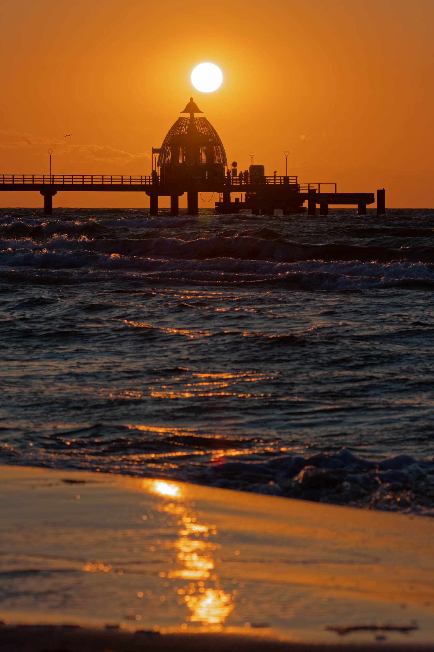 Das Bild zeigt einen atemberaubenden Sonnenuntergang über dem Meer. Die Sonne steht tief am Horizont und wirft ein warmes, goldenes Licht über die Szene. Im Vordergrund erstreckt sich ein Steg, der sich elegant über das Wasser erhebt. Die Silhouette des Stegs, mit seinen charakteristischen Kuppeln und Geländern, bildet einen starken Kontrast zum leuchtenden Himmel. Das Wasser ist leicht aufgewühlt und reflektiert das Licht der untergehenden Sonne. Der Strand im Vordergrund ist dunkel und spiegelt ebenfalls das warme Licht wider. Die Szene vermittelt eine friedliche und ruhige Atmosphäre.