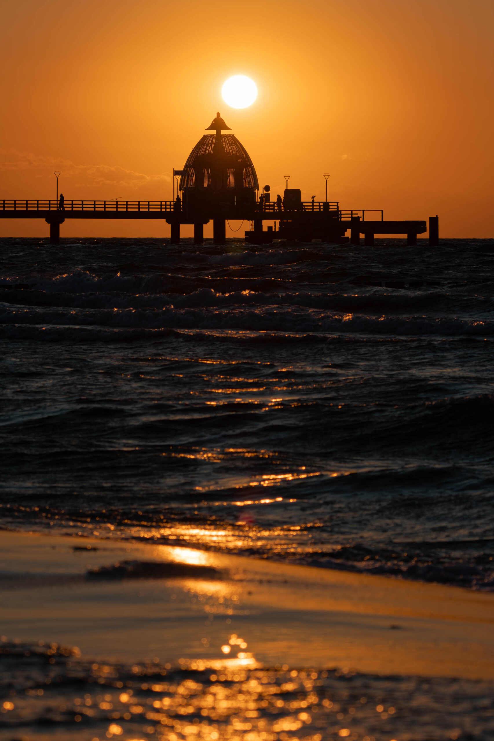 Das Bild zeigt einen atemberaubenden Sonnenuntergang über der Ostsee, aufgenommen im Sommer 2025 in Zingst. Im Vordergrund erstreckt sich ein langer Steg, der sich ins Wasser erstreckt. Die Silhouette des Stegs und der darauf befindlichen Gebäude wird gegen den leuchtenden, orangefarbenen Himmel abgezeichnet. Die Sonne, fast vollständig im Meer versunken, erzeugt einen goldenen Schein auf der Wasseroberfläche und dem feuchten Sandstrand. Einige Personen sind auf dem Steg zu erkennen, die sich in der Silhouette des Bauwerks verlieren. Die Szene vermittelt eine friedliche und ruhige Atmosphäre.