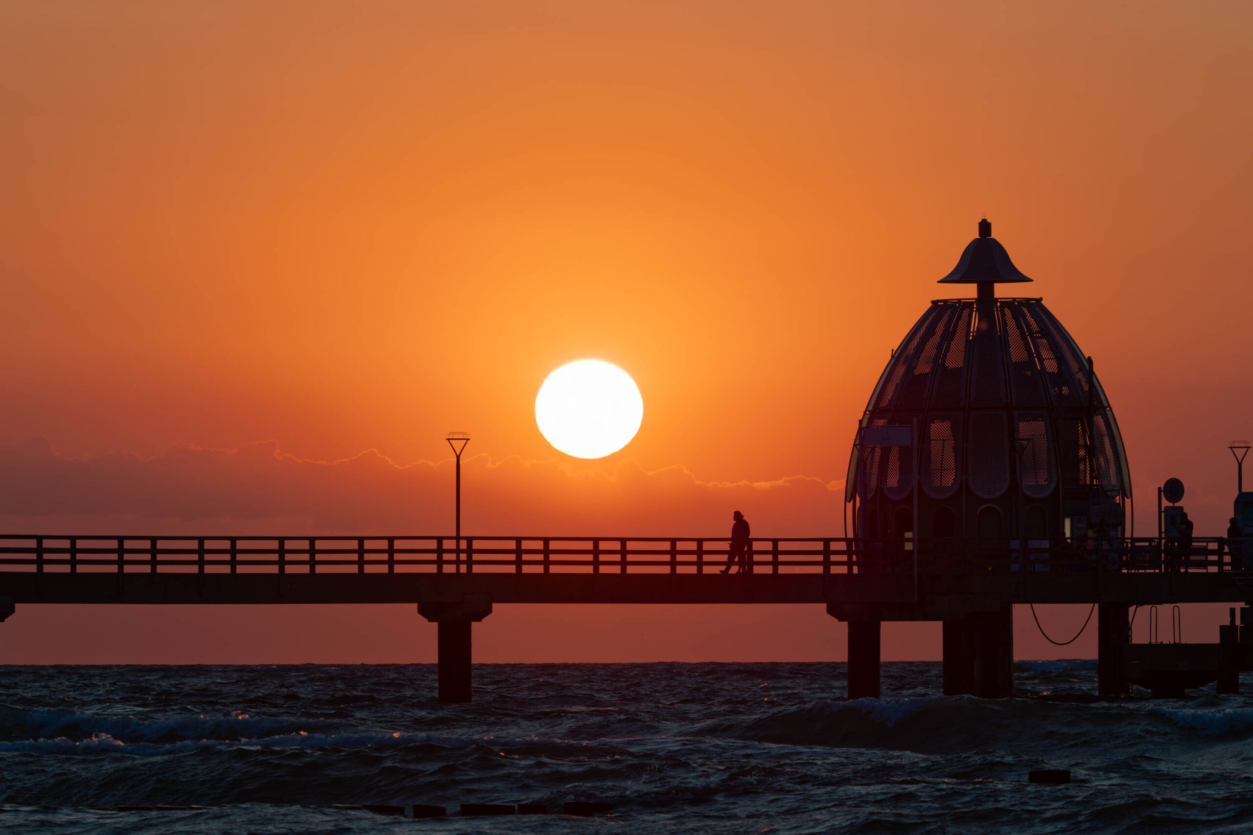 Das Bild zeigt einen atemberaubenden Sonnenuntergang über dem Seebrückengebäude in Zingst, Deutschland. Die Sonne, ein leuchtender Kreis, steht tief am Horizont und taucht den Himmel in warme Orange- und Rottöne. Das Seebrückengebäude, ein markantes Bauwerk mit einer Kuppel, steht im Vordergrund und ist silhouettiert gegen das helle Licht der untergehenden Sonne. Auf der Brücke sind einige Personen zu sehen, die ebenfalls silhouettiert sind. Das Wasser ist leicht bewegt und reflektiert das Licht des Himmels. Die Szene vermittelt eine friedliche und entspannte Atmosphäre.