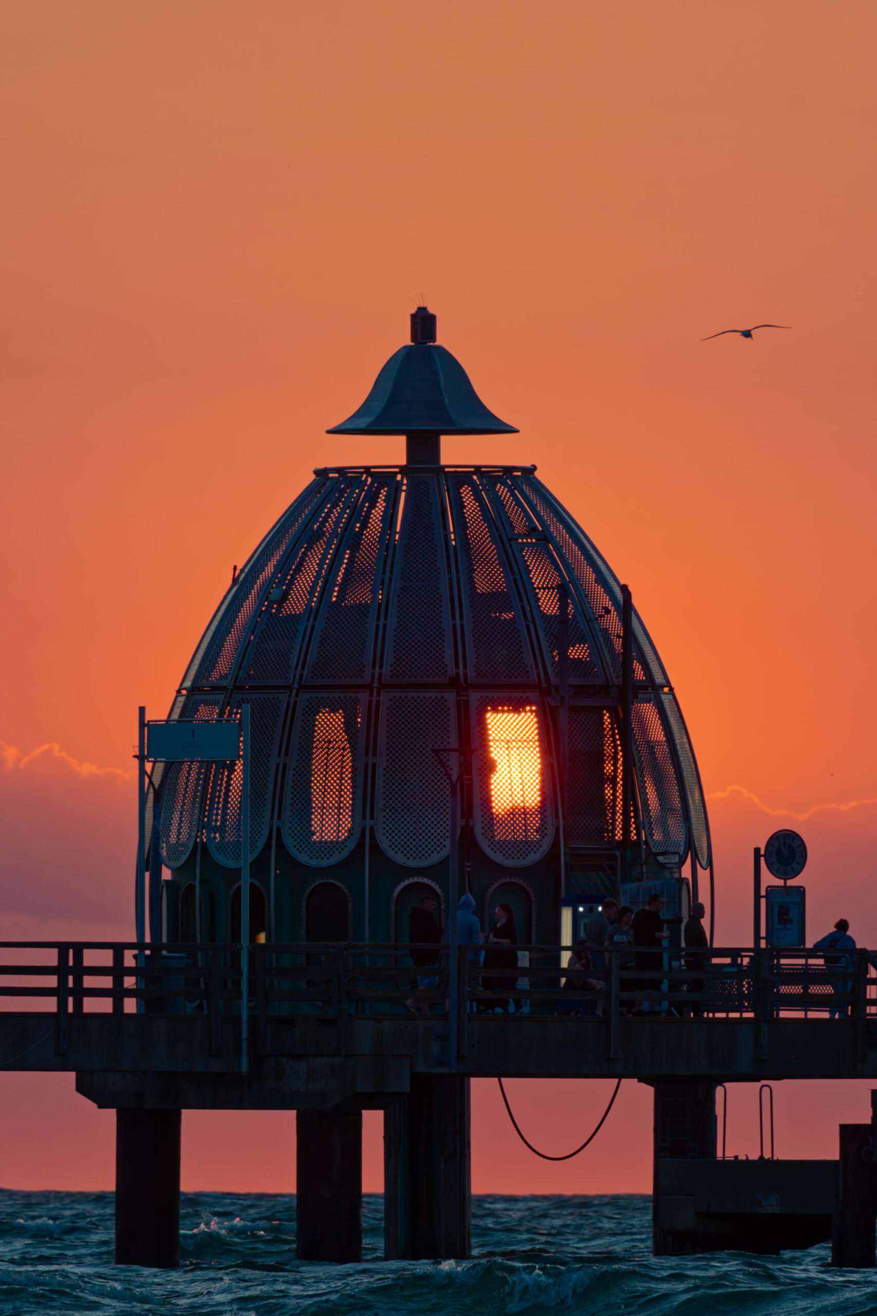 Das Foto zeigt den Zingster Seebrückenturm bei Sonnenuntergang. Der Himmel ist in warmen Orange- und Rottönen gehalten, was eine stimmungsvolle Atmosphäre schafft. Der Seebrückenturm, ein markantes Bauwerk, steht auf einem hölzernen Steg, der sich ins Meer erstreckt. Das Licht im Inneren des Turms erhellt die filigranen Gitterstrukturen. Im Hintergrund sind einige Personen auf dem Steg erkennbar. Ein einzelner Vogel fliegt am Himmel.