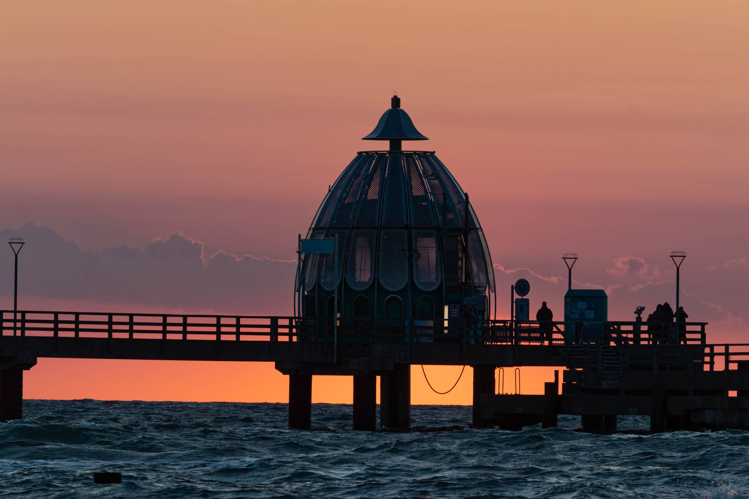 Die Aufnahme zeigt eine hölzerne Seebrücke, die sich vor einem leuchtenden, orangefarbenen Himmel erstreckt. Die Brücke ist im Vordergrund als Silhouette dargestellt, was die dramatische Wirkung des Sonnenuntergangs verstärkt. Das markante Kuppelgebäude der Seebrücke ist deutlich erkennbar. Das Wasser ist leicht aufgewühlt und spiegelt die Farben des Himmels wider. Im Hintergrund sind einige Personen auf der Brücke zu erkennen. Die Aufnahme vermittelt eine ruhige und friedliche Atmosphäre.
