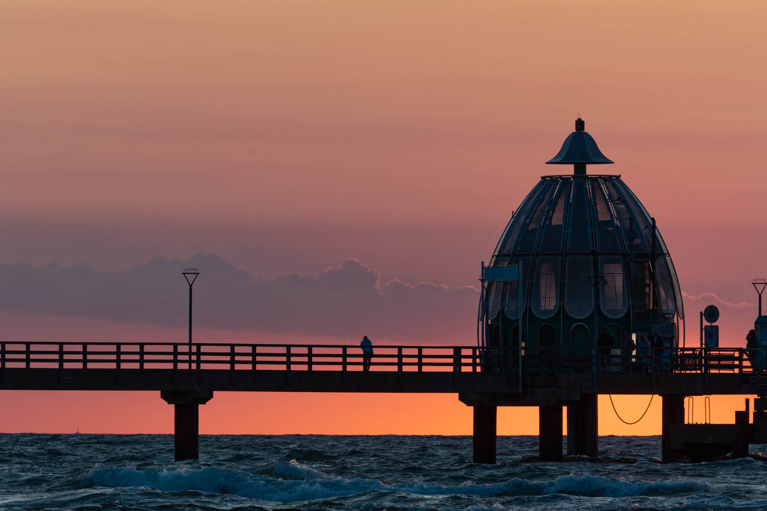Das Foto zeigt den Zingster Seebrückens bei Sonnenuntergang. Der Himmel ist in warmen Orange- und Rottönen gehalten, die sich im Wasser spiegeln. Die Seebrücke, mit ihrem markanten Kuppelbau, ist im Vordergrund als Silhouette erkennbar. Einige Personen sind auf der Brücke zu sehen. Das Meer ist leicht aufgewühlt, mit kleinen Wellen, die an die Küste rollen. Im Hintergrund sind die Küstenlinien und einige Gebäude erkennbar. Die Szene vermittelt eine ruhige und friedliche Atmosphäre.