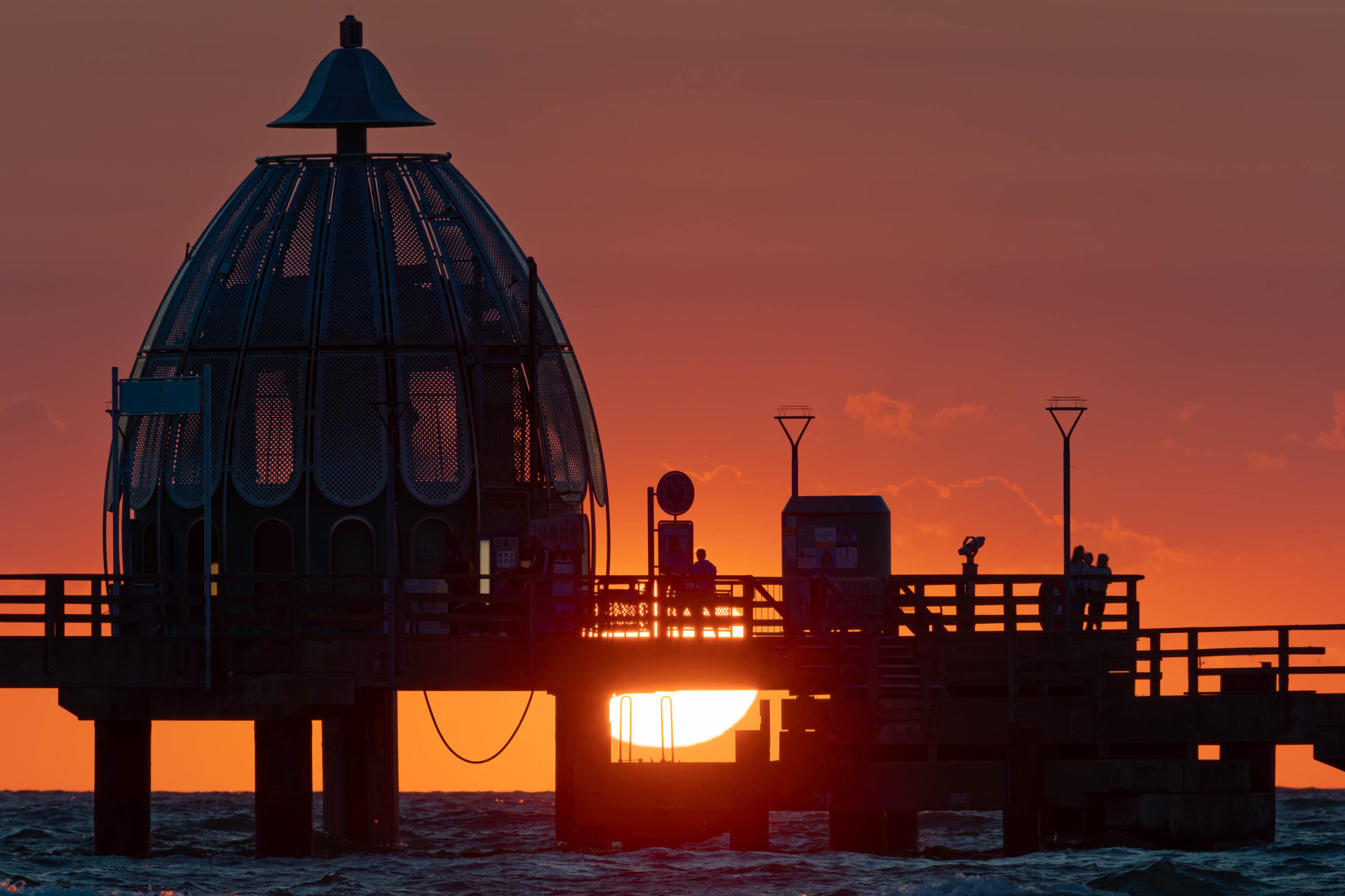 Die Aufnahme zeigt einen Pier bei Sonnenuntergang. Der Pier ist im Vordergrund und erhebt sich aus dem Wasser. Der Himmel ist in leuchtenden Orange- und Rottönen gehalten, was die Szene dramatisch und stimmungsvoll macht. Die Silhouette des Piers, einschließlich seiner markanten Kuppel, ist deutlich erkennbar. Mehrere Personen befinden sich auf dem Pier und scheinen den Sonnenuntergang zu beobachten. Das Wasser ist ruhig und spiegelt die Farben des Himmels wider. Die Aufnahme ist im Stil einer Silhouettenaufnahme gehalten, wobei der Fokus auf den Konturen des Piers und der Personen liegt. Die Aufnahme vermittelt ein Gefühl von Ruhe, Frieden und Nostalgie.