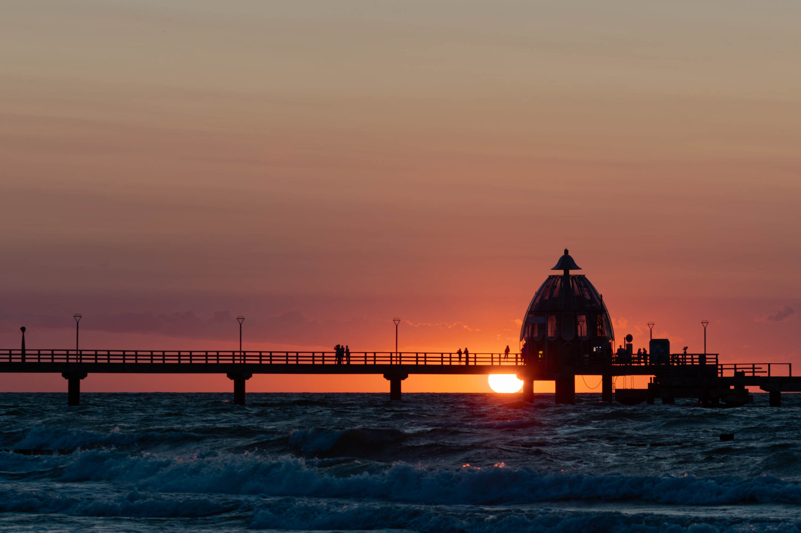 Die Aufnahme zeigt eine Seebrücke vor dem Hintergrund eines dramatischen Sonnenuntergangs. Die Brücke erstreckt sich horizontal über das Bild und wird von mehreren Pfeilern gestützt, die aus dem Wasser ragen. Die Silhouette der Brücke und der Personen darauf wird vor dem leuchtenden orange-roten Himmel deutlich hervorgehoben. Das Wasser ist leicht aufgewühlt und reflektiert das Licht des Sonnenuntergangs. Die Aufnahme vermittelt eine friedliche und ruhige Atmosphäre.