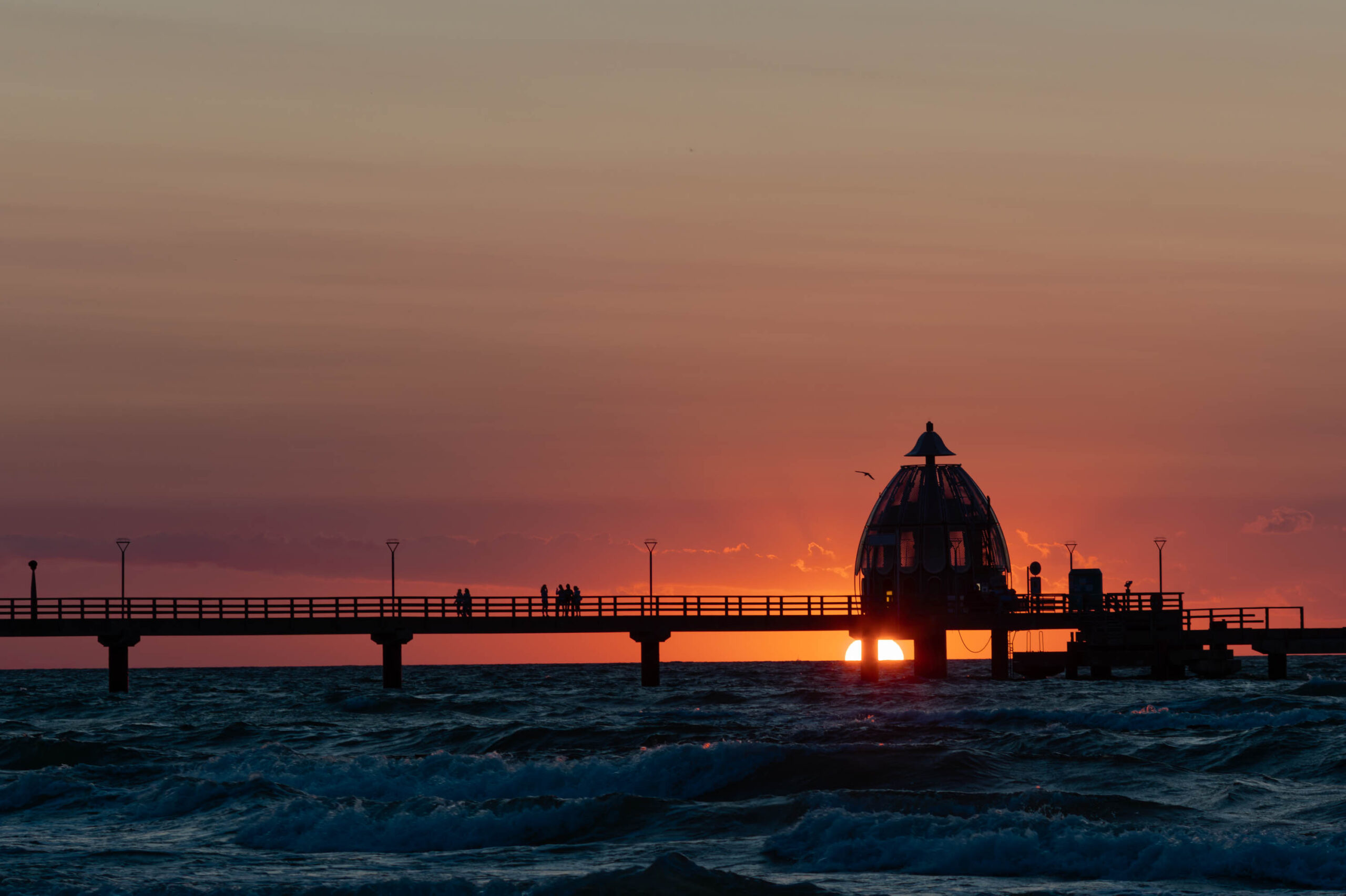 Das Bild zeigt einen atemberaubenden Sonnenuntergang über dem Meer, wobei ein hölzerner Pier im Vordergrund dominiert. Der Himmel ist in leuchtenden Rottönen und Orangetönen gefärbt, die sich im Wasser spiegeln. Der Pier, der auf Stelzen errichtet ist, erstreckt sich weit ins Meer hinein und ist von mehreren Personen belebt, die sich entspannt aufhalten. Die Silhouette des Piers und der Menschen bildet einen starken Kontrast zum farbenprächtigen Himmel. Die Wellen sind leicht bewegt und tragen zur dynamischen Atmosphäre des Bildes bei. Es ist ein ruhiger und friedlicher Moment, eingefangen in einem wunderschönen Sonnenuntergang.