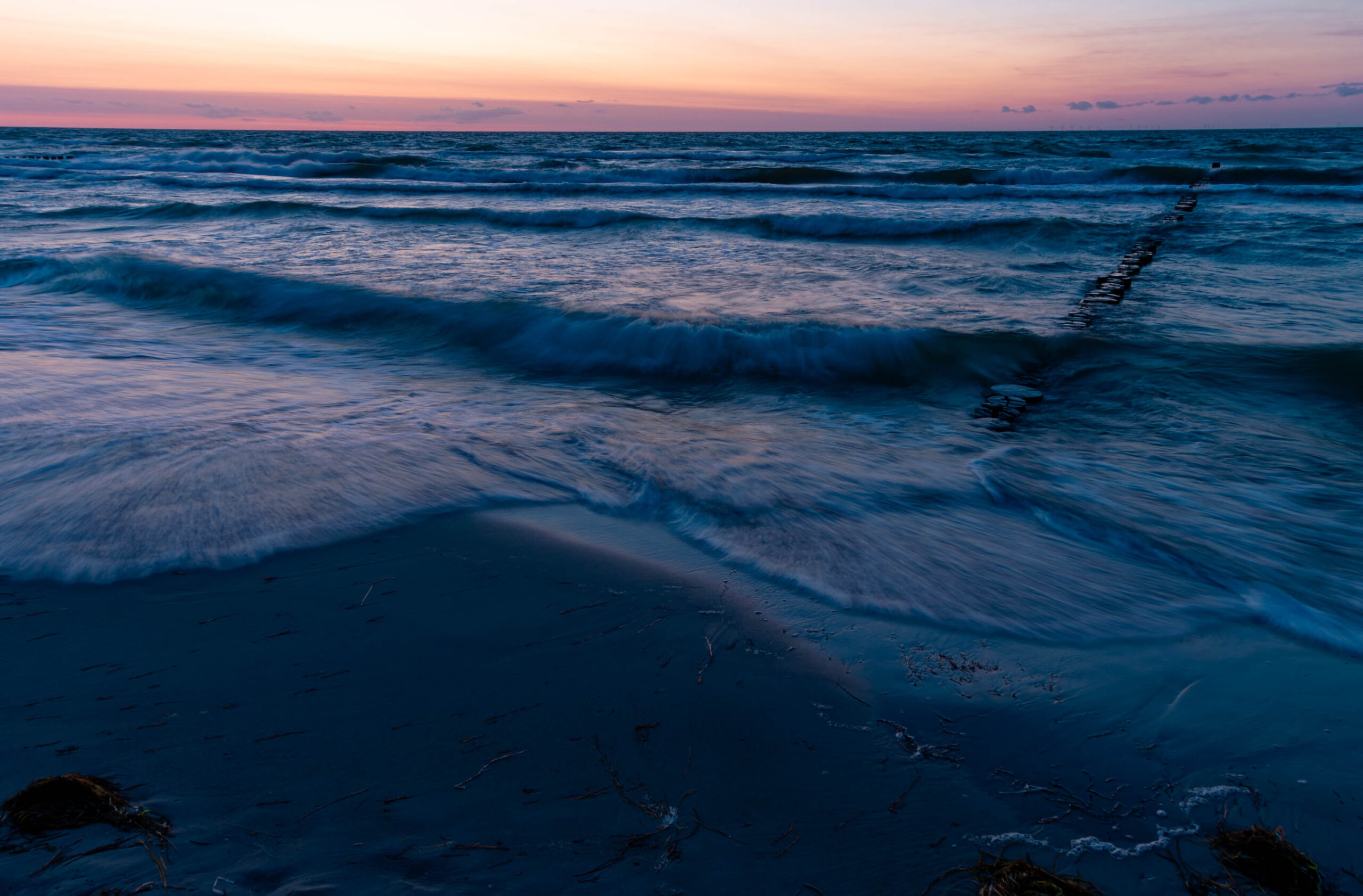 Das Foto zeigt einen Strand bei Zingst, aufgenommen im Sommer 2025. Das Licht der untergehenden Sonne färbt den Himmel in zarten Rosatönen und Violett. Die Wellen brechen sanft auf den dunklen Sandstrand und hinterlassen eine Spur aus Schaum. Im Vordergrund sind Treibholz und Seegras verstreut. Ein hölzerner Steg erstreckt sich in die Ferne und führt ins Meer. Die Szene vermittelt eine ruhige und friedliche Atmosphäre.