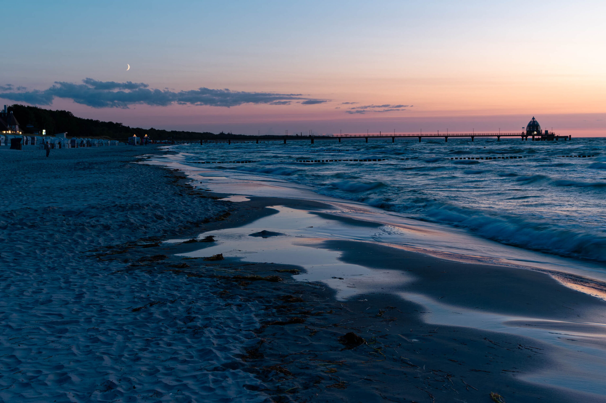 Die Aufnahme zeigt einen ruhigen Sonnenuntergang an der Küste von Zingst, Deutschland. Der Himmel ist in sanften Rosa- und Lilatönen gehalten, die sich im feuchten Sand spiegeln. Ein hölzerner Steg, vermutlich der Zingster Steg, erstreckt sich weit ins Meer. Im Hintergrund sind vereinzelte Personen zu sehen, die den Sonnenuntergang genießen. Die Szene vermittelt eine friedliche und entspannte Atmosphäre.