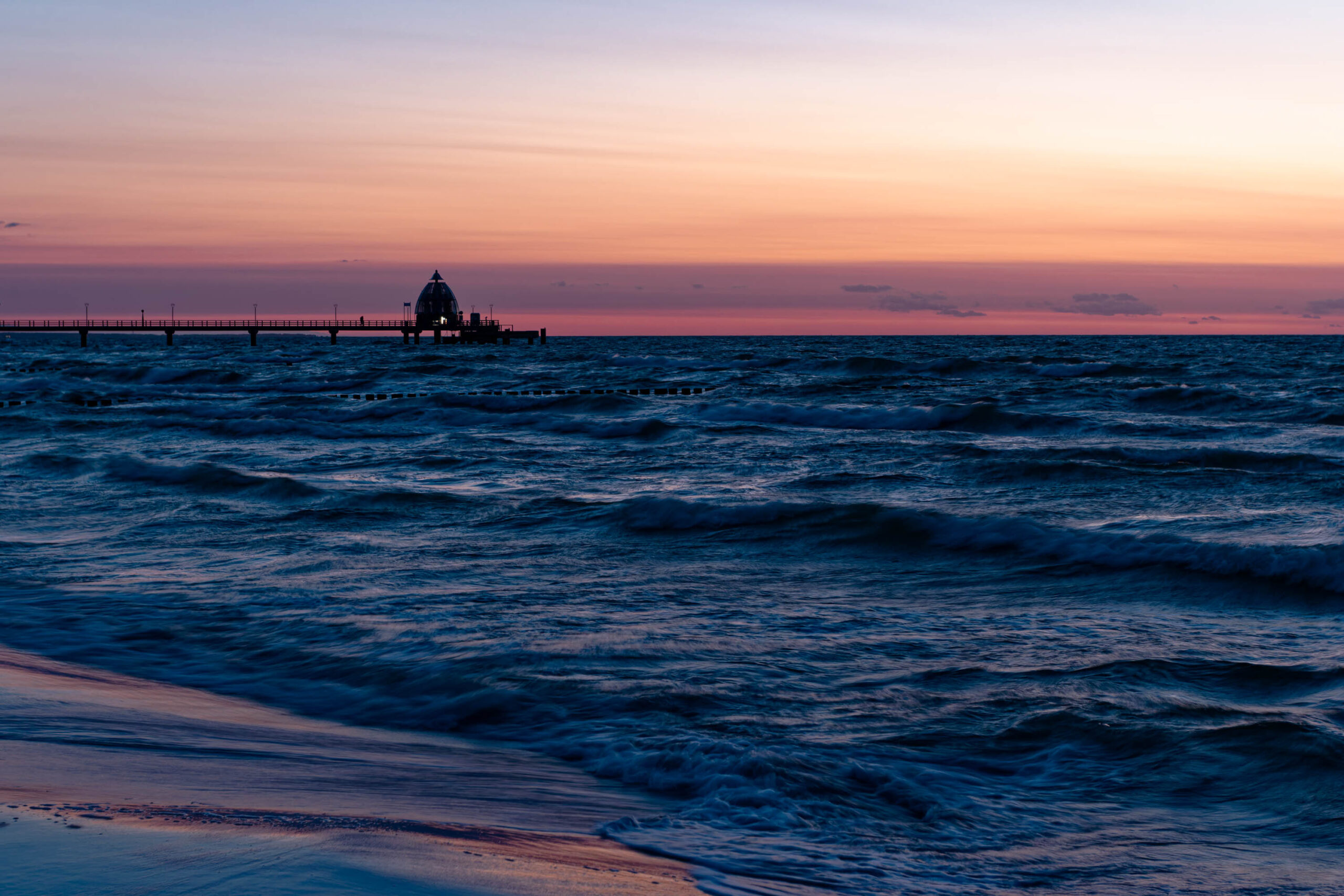 Das Foto zeigt einen Steg, der sich in das dunkle, wellige Meer erstreckt. Der Himmel ist in einem spektakulären Sonnenuntergang in warmen Farbtönen von Rosa und Orange gefärbt. Der Strand im Vordergrund ist sandig und leicht feucht. Die Szene vermittelt eine ruhige und friedliche Atmosphäre, unterbrochen durch die Bewegung des Wassers.