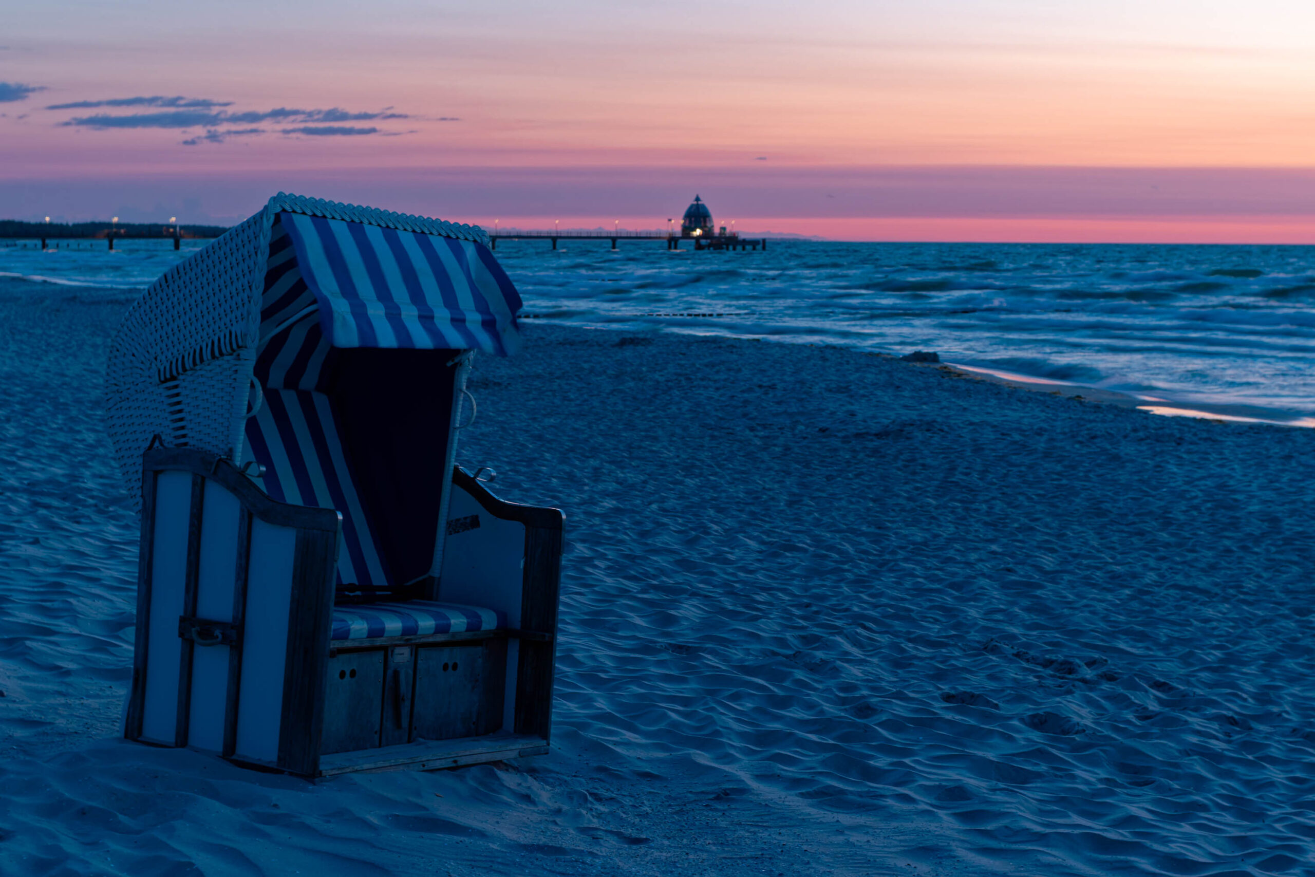 Das Foto zeigt einen typischen Strandkorb auf dem Sandstrand von Zingst. Die Szene ist in ein sanftes, warmes Licht getaucht, das von einem Sonnenuntergang herrührt. Der Himmel ist in zarten Rosa- und Lilatönen gehalten, was eine ruhige und friedliche Atmosphäre schafft. Im Hintergrund ist ein hölzerner Steg und das Ostseewasser zu sehen, das in sanften Wellen an den Strand rollt. Die Szene vermittelt ein Gefühl von Sommer, Ruhe und Erholung.