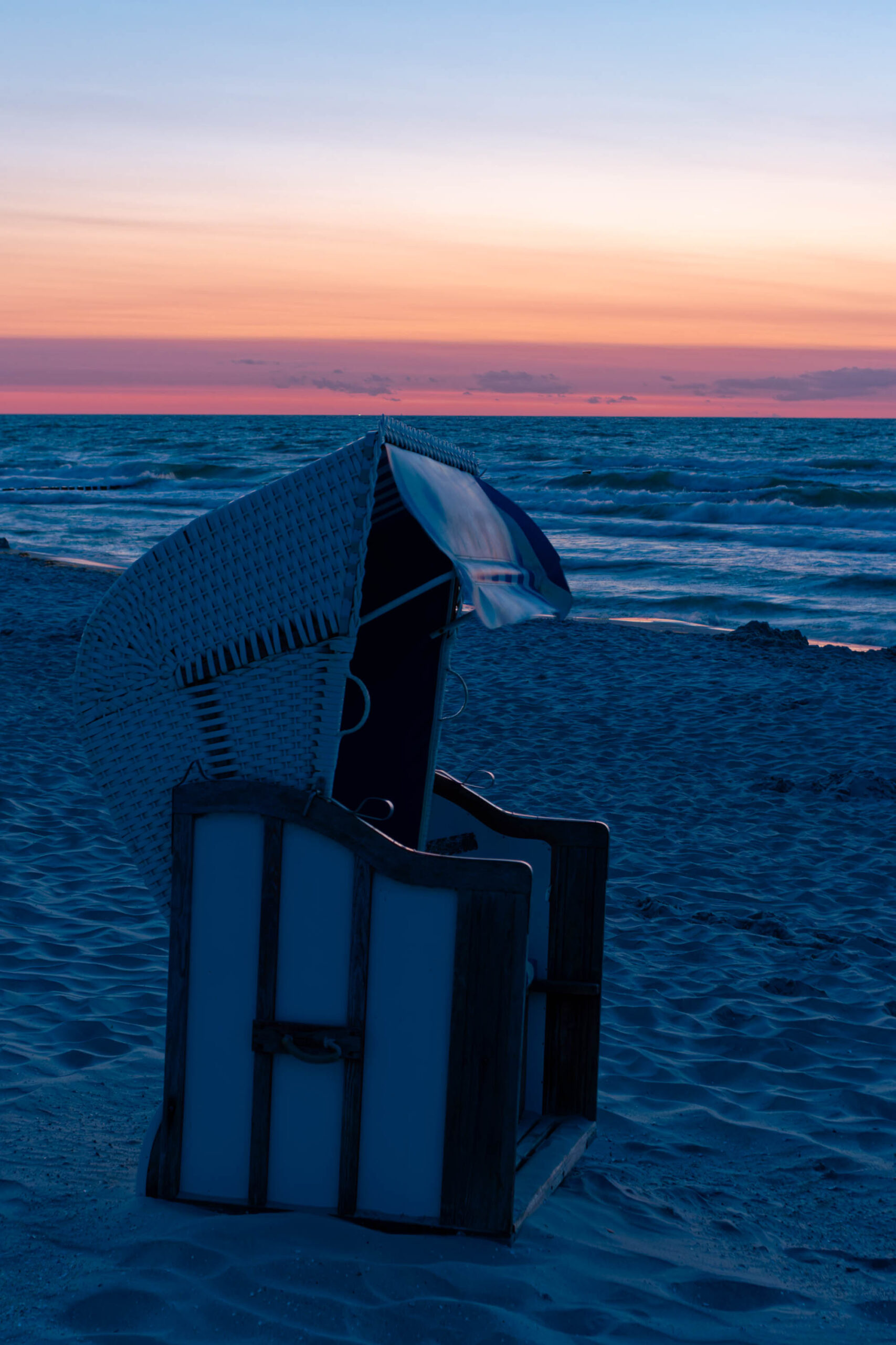 Ein Strandkorb steht im Vordergrund vor einem Sonnenuntergang am Strand von Zingst. Der Himmel ist in warmen Farben wie Orange und Rosa gehalten, die sich im feuchten Sand spiegeln. Die Wellen brechen sanft am Ufer. Die Szene vermittelt eine friedliche und entspannte Atmosphäre.