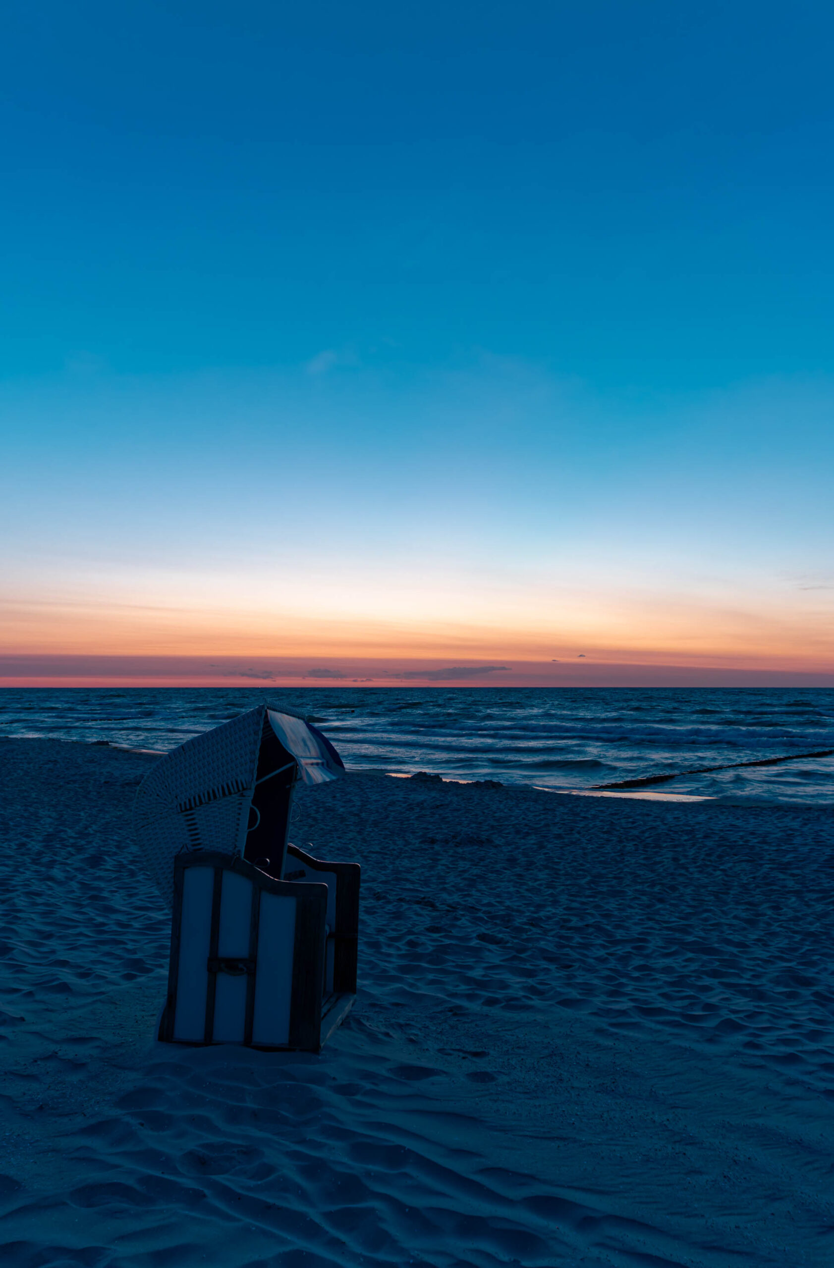 Das Bild zeigt einen typischen Strandkorb, der auf einem Sandstrand steht. Der Himmel ist in warmen Orange- und Blautönen gehalten, was auf einen Sonnenuntergang hindeutet. Das Wasser ist ruhig und spiegelt die Farben des Himmels wider. Der Sand ist feucht und weist leichte Wellenmuster auf. Die Szene vermittelt eine friedliche und entspannte Atmosphäre. Der Strandkorb ist ein charakteristisches Element der Ostseeküste und verleiht dem Bild einen regionalen Bezug.