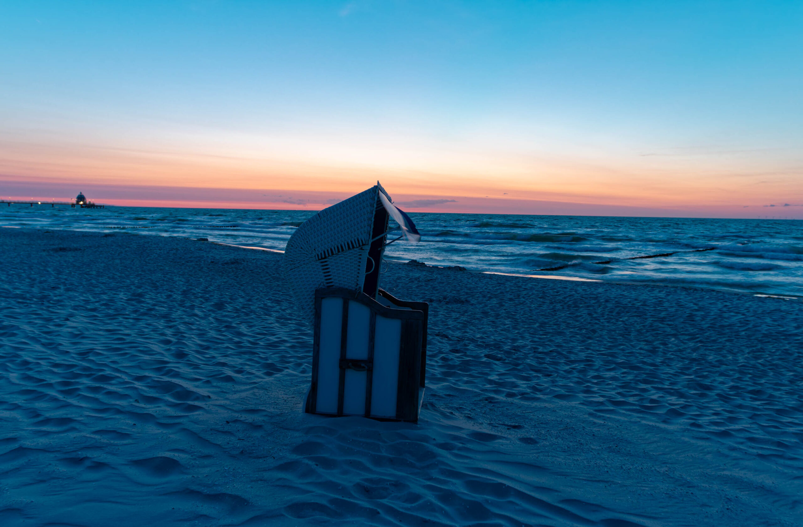 Das Bild zeigt einen typischen Strandkorb auf einem Sandstrand in Zingst, aufgenommen im Sommer 2025. Der Strand ist feucht und reflektiert das Licht des Sonnenuntergangs. Im Hintergrund ist eine Seebrücke erkennbar, die die Weite des Meeres betont. Die Szene vermittelt eine ruhige und friedliche Atmosphäre.