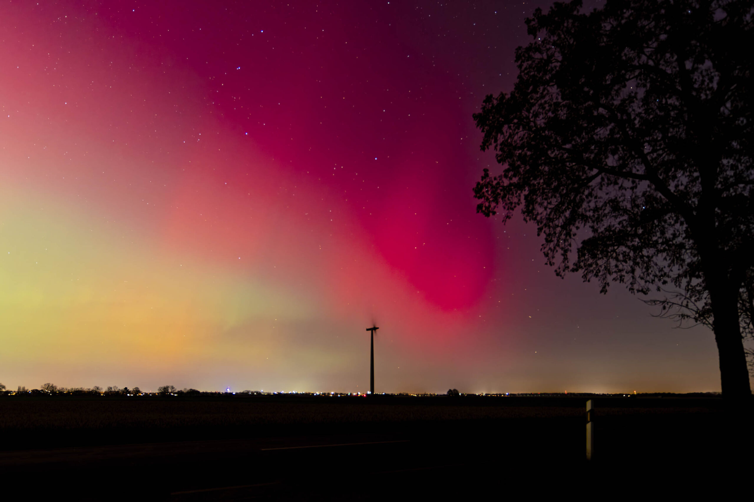 Ein atemberaubendes Polarlicht erhellt den Nachthimmel über einer ländlichen Landschaft. Die leuchtenden Farben des Polarlichts kontrastieren mit den dunklen Silhouetten der Bäume und der Stadt am Horizont. Ein einzelner Strommast steht als stummer Zeuge dieses spektakulären Himmelsphänomens.